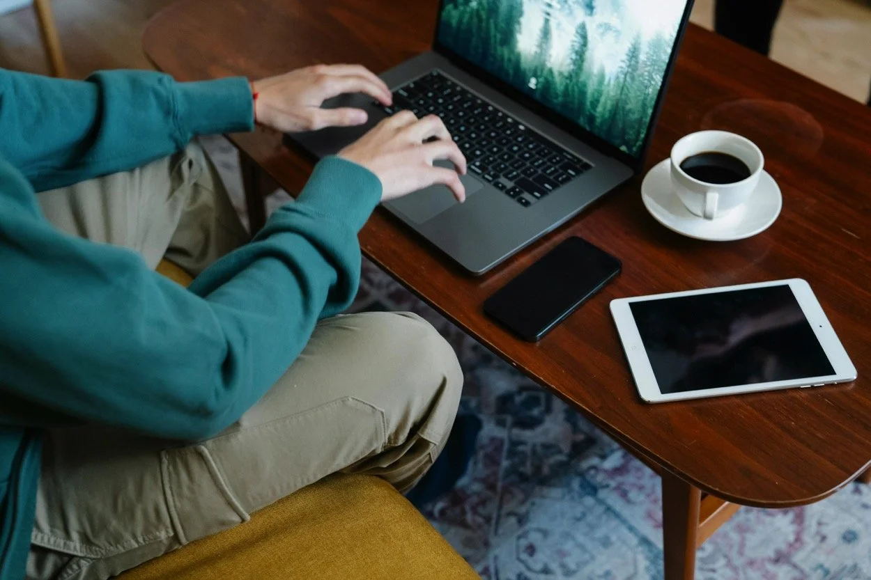 Person working on a laptop with a smartphone and tablet on a wooden table, a cup of coffee nearby.