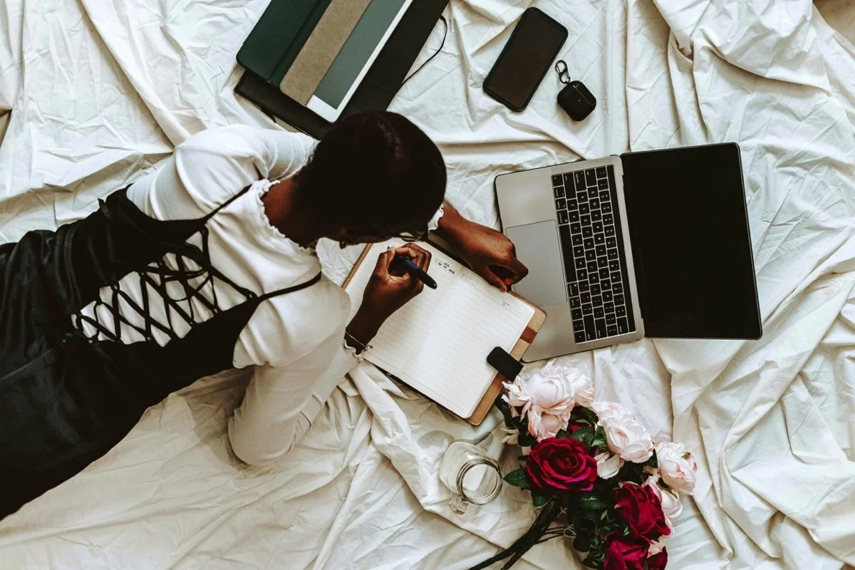 Person lying on a bed with a laptop, notebook, smartphone, wireless earbuds, and flowers around them.