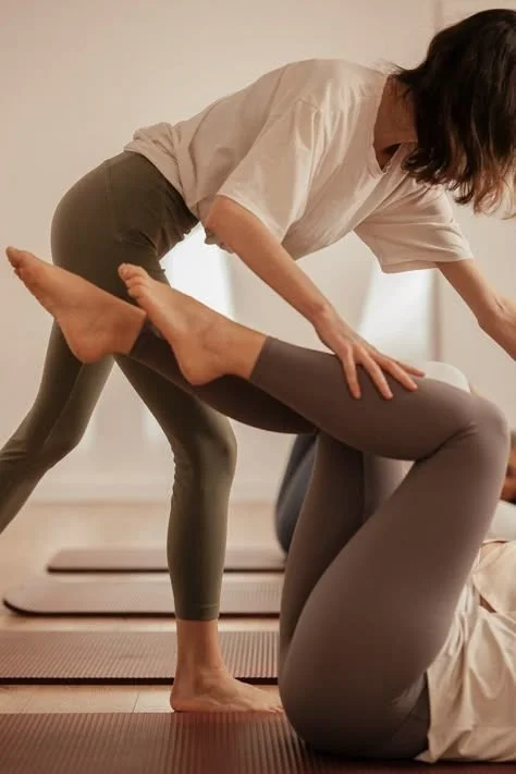 Two women practicing yoga, one assisting the other during a pose in a studio with yoga mats.