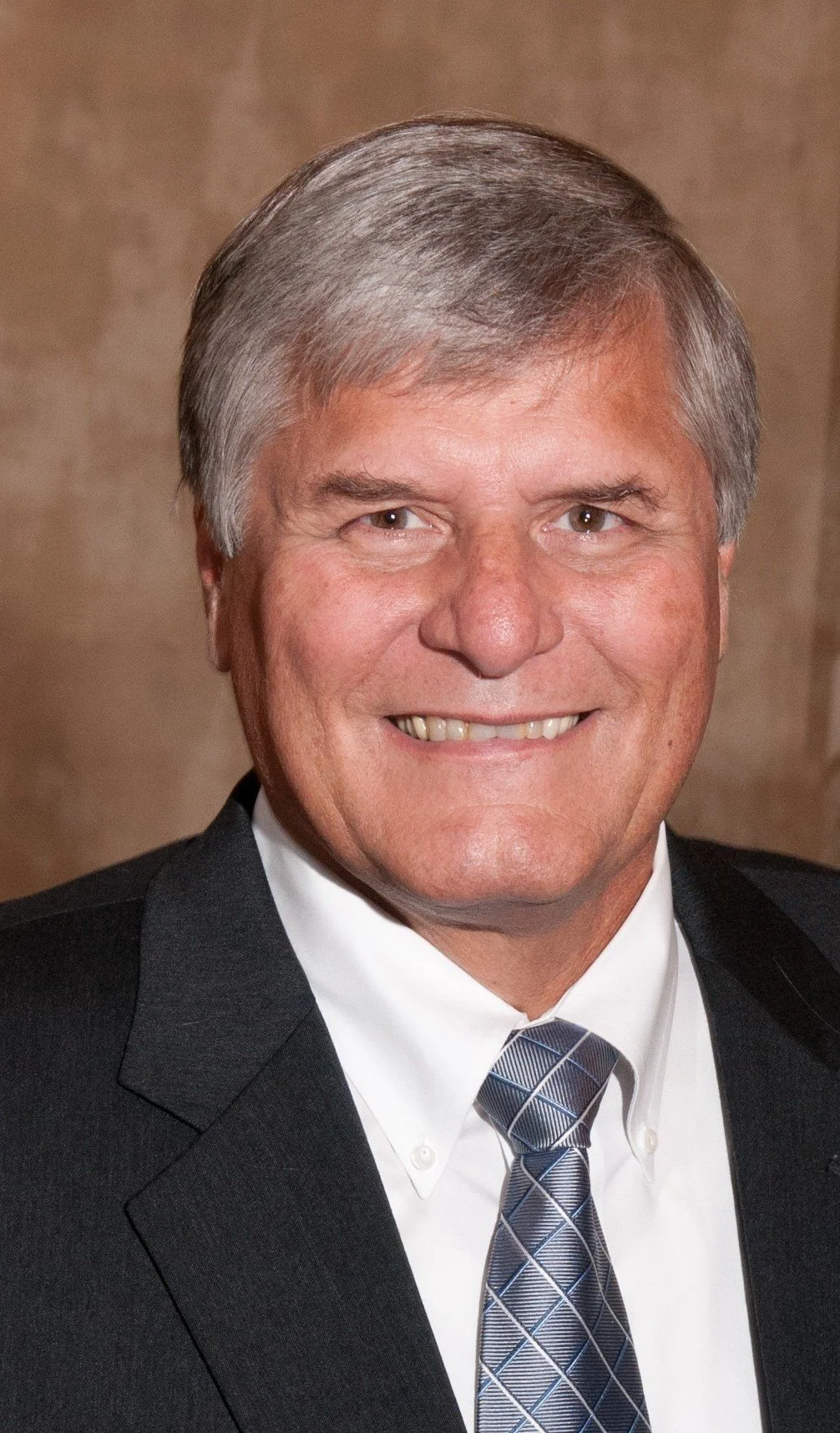 A middle-aged man with gray hair, wearing a black suit and a patterned blue tie, smiling at the camera.