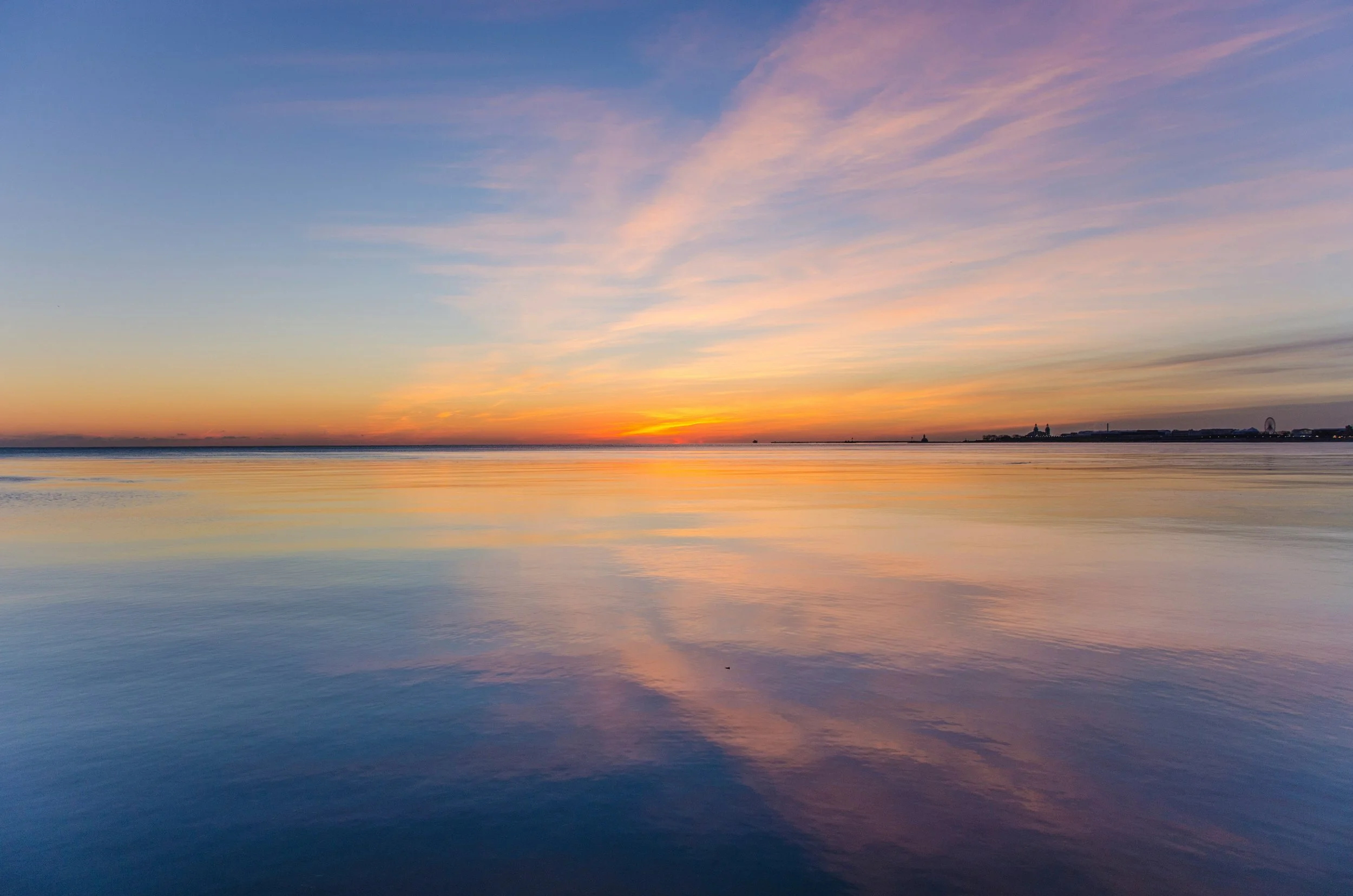 A calm body of water reflecting a colorful sunset sky with pink, purple, and orange hues, distant land with silhouettes of buildings and a Ferris wheel on the horizon.
