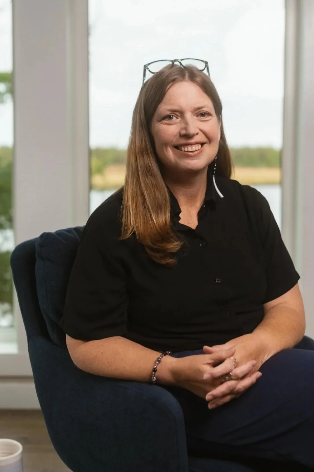 Portrait shot of a woman with medium length light brown hair, glasses on her head, dressed all in black sitting in a chair with her arms crossed in front of her. There is a blurry landscape of a lake and treeline in the background.