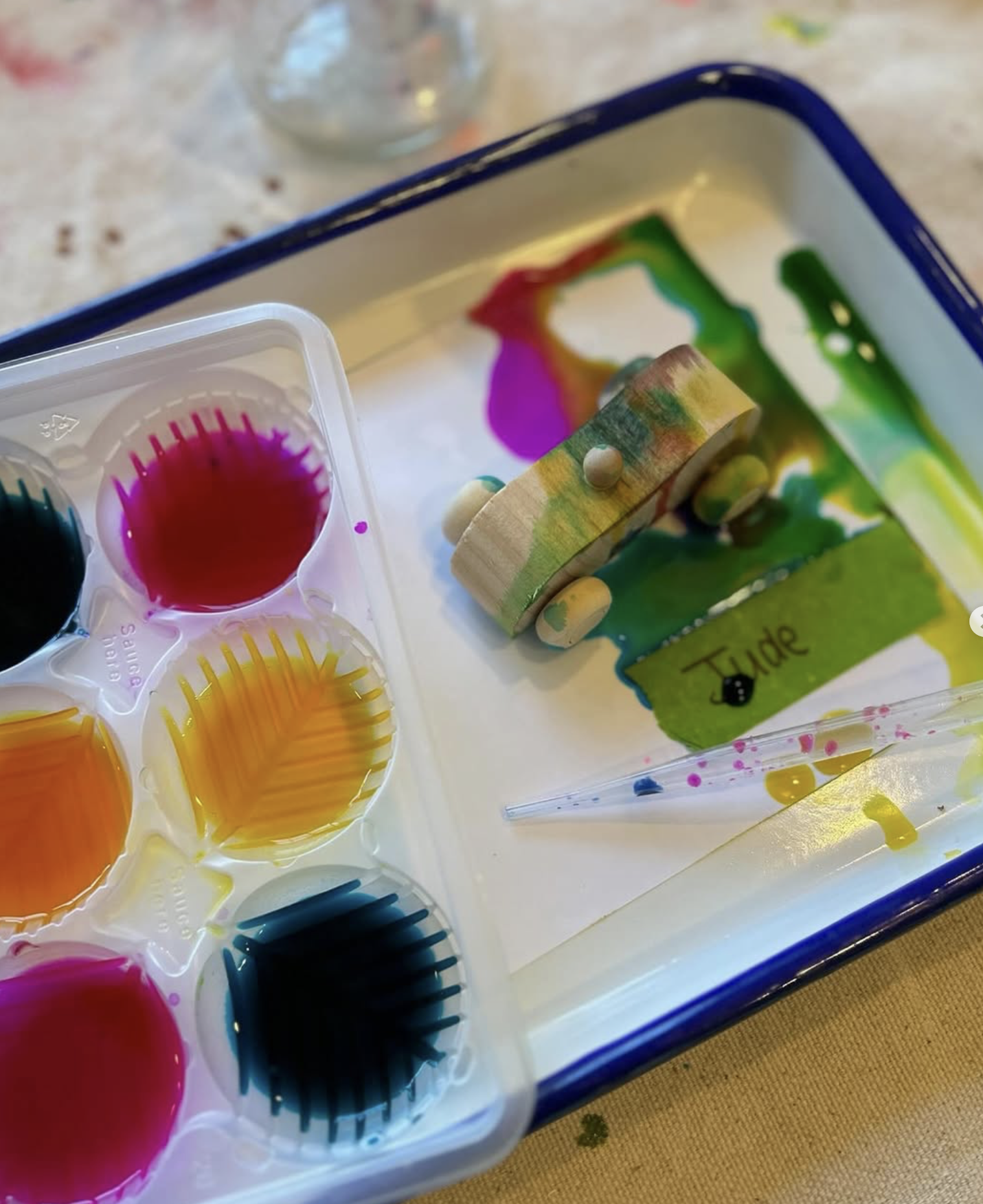 Paint palette with vibrant colors, a small wooden toy with wheels, and a brush, all on a tray.
