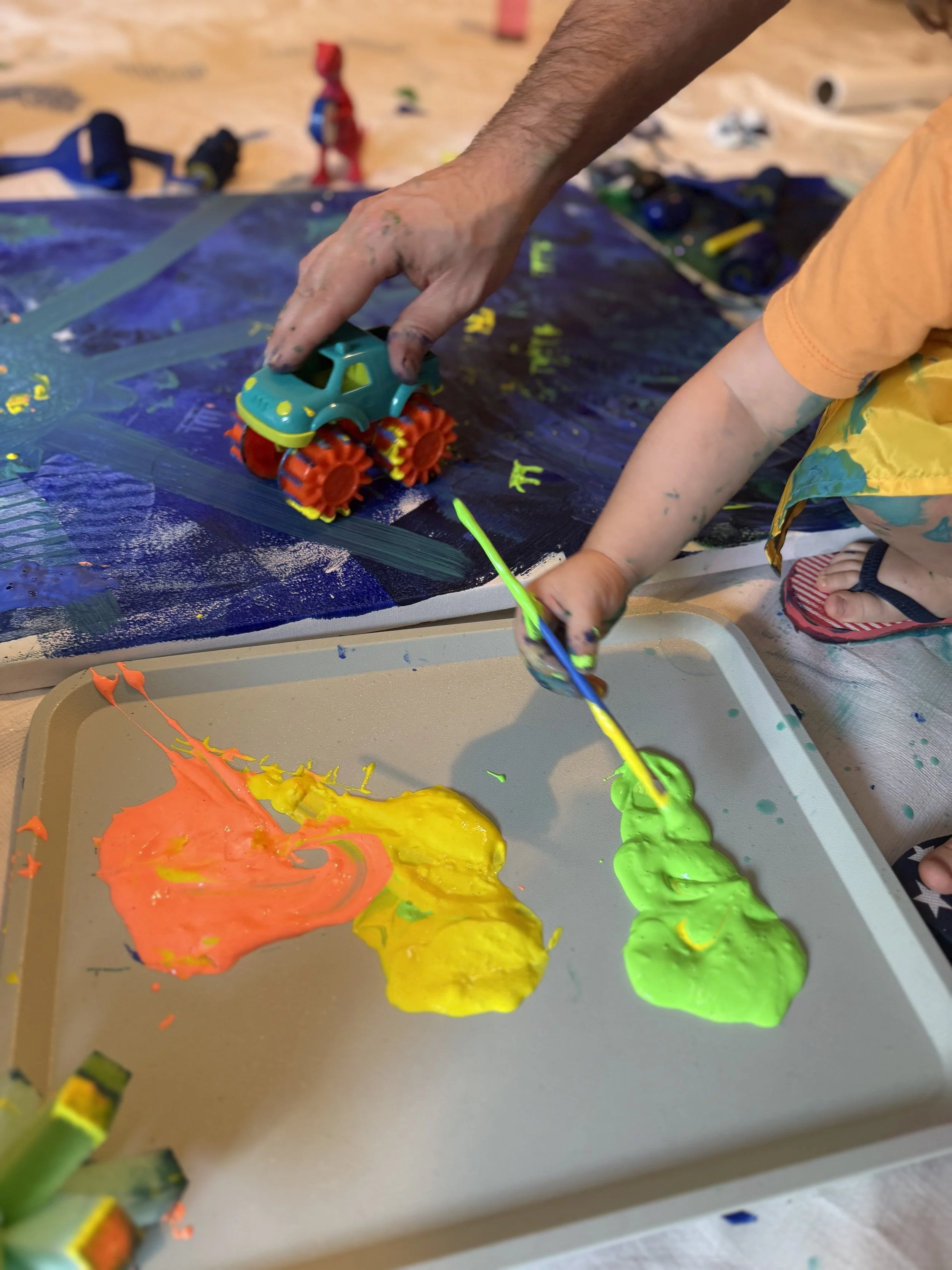 Child painting on a large fabric-covered outdoor board with colorful paint, standing on grass near a tree stump with art supplies, in a backyard with trees and a white fence in the background.