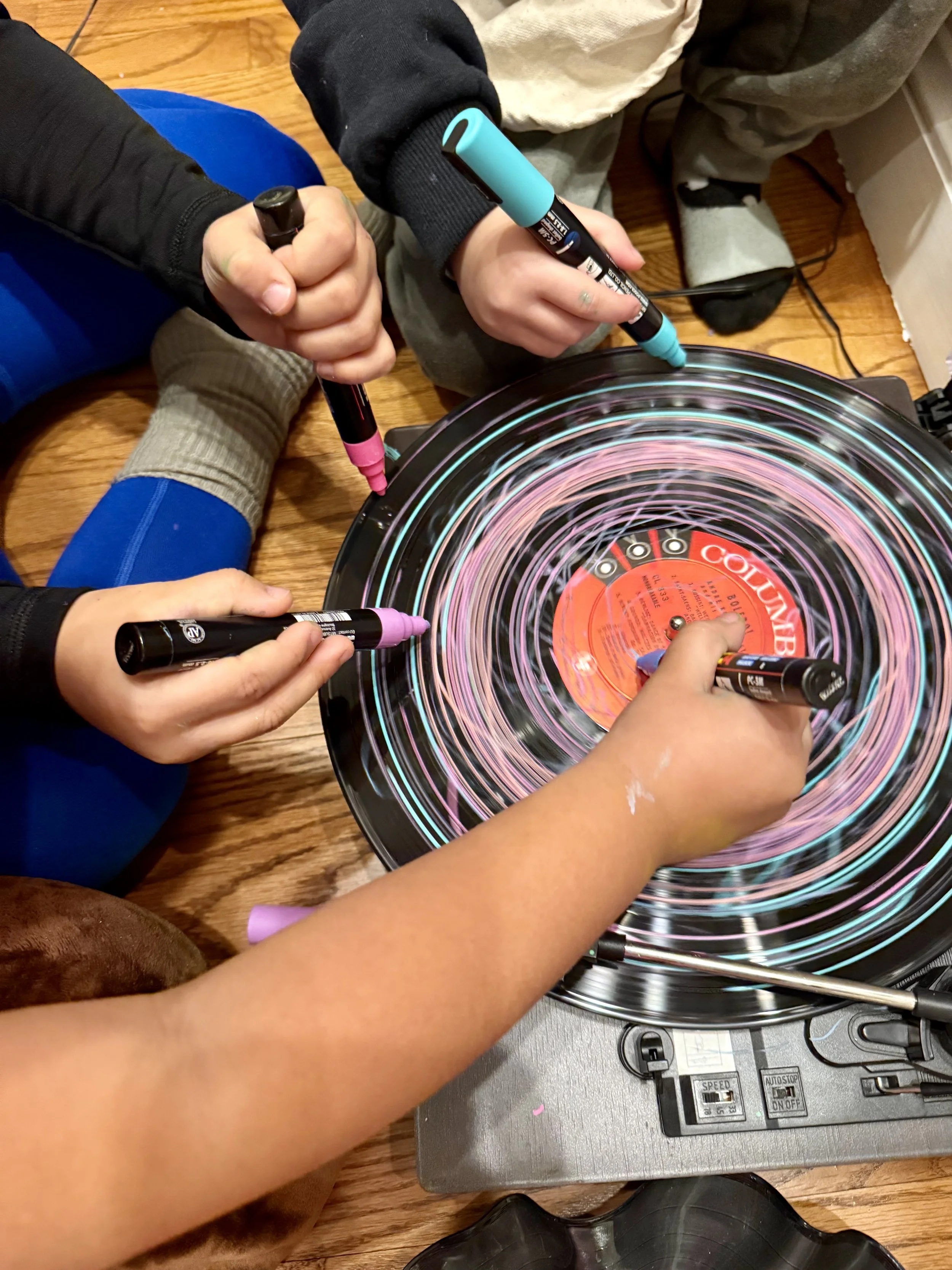 Three children drawing colorful spiral patterns on a spinning vinyl record with markers.