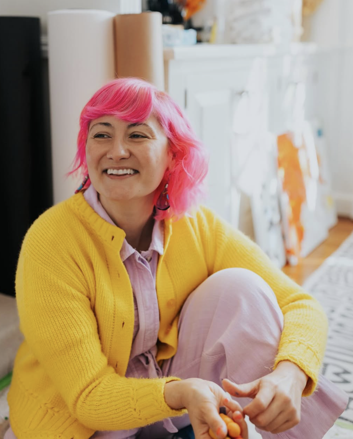 A person with pink hair, wearing a yellow cardigan and pink pants, sitting indoors and smiling.