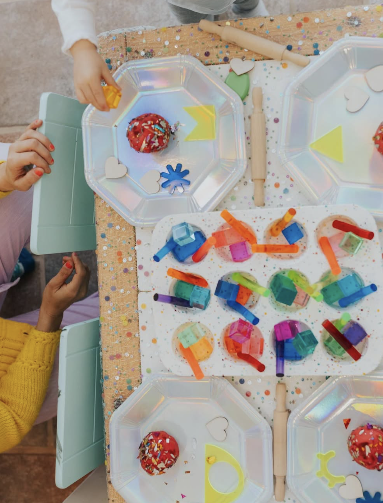 Children are sitting at a table with colorful craft supplies, including small transparent blocks, straws, and decorated cupcakes with sprinkles, surrounded by festive decorations.