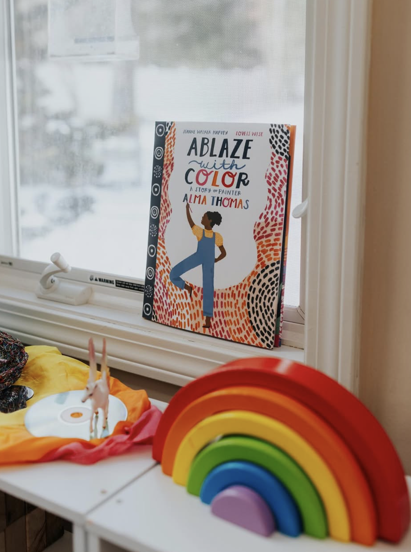 A window sill with a colorful children's book titled 'A Blaze with Color,' a rainbow stacking toy, a small unicorn figurine on a fabric surface, and a snowy street scene outside the window.