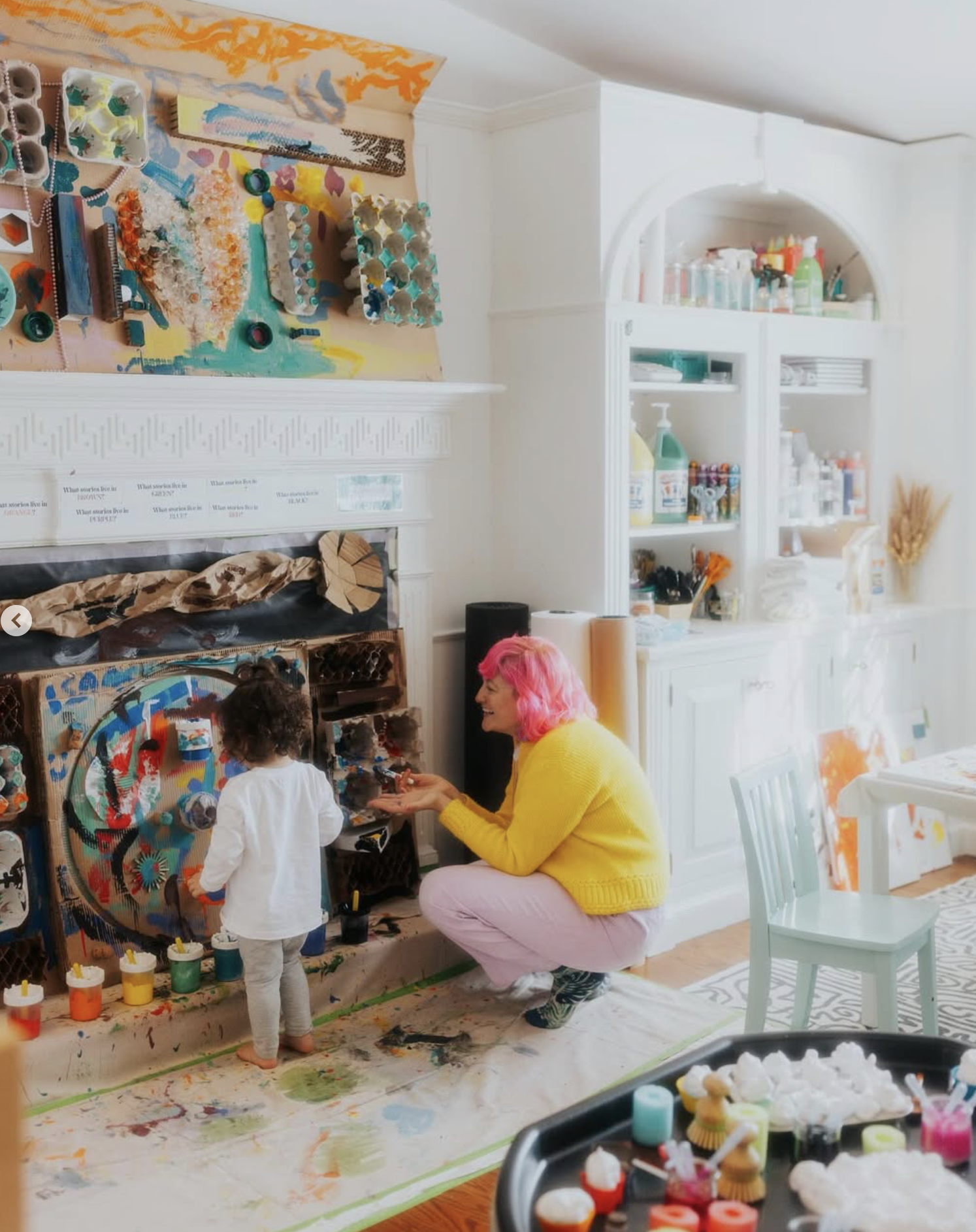 A person with pink hair and a yellow sweater squats next to a small child, both engaging in a colorful art activity in a well-lit room with shelves of art supplies and a table.
