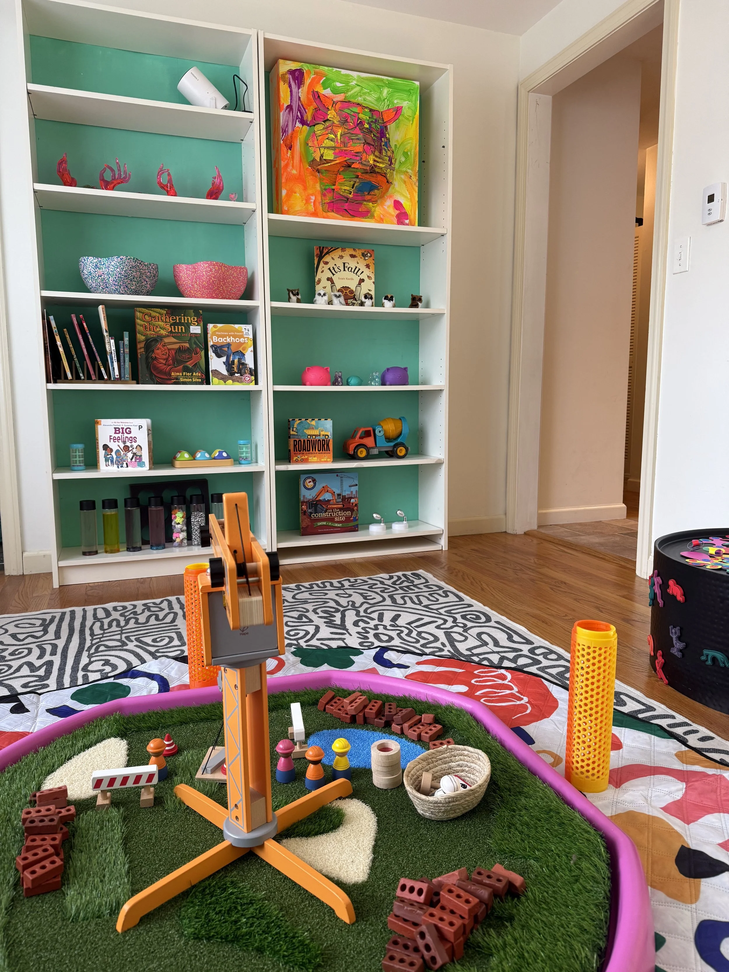 Kids' indoor play area with a colorful, circular mini golf setup on a green artificial turf rug, a toy construction crane, stacking toys, and a basket of mini golf balls. In the background, there is a tall white bookshelf filled with books, toys, and colorful decorations, including a bright abstract painting of a tiger on the top shelf.
