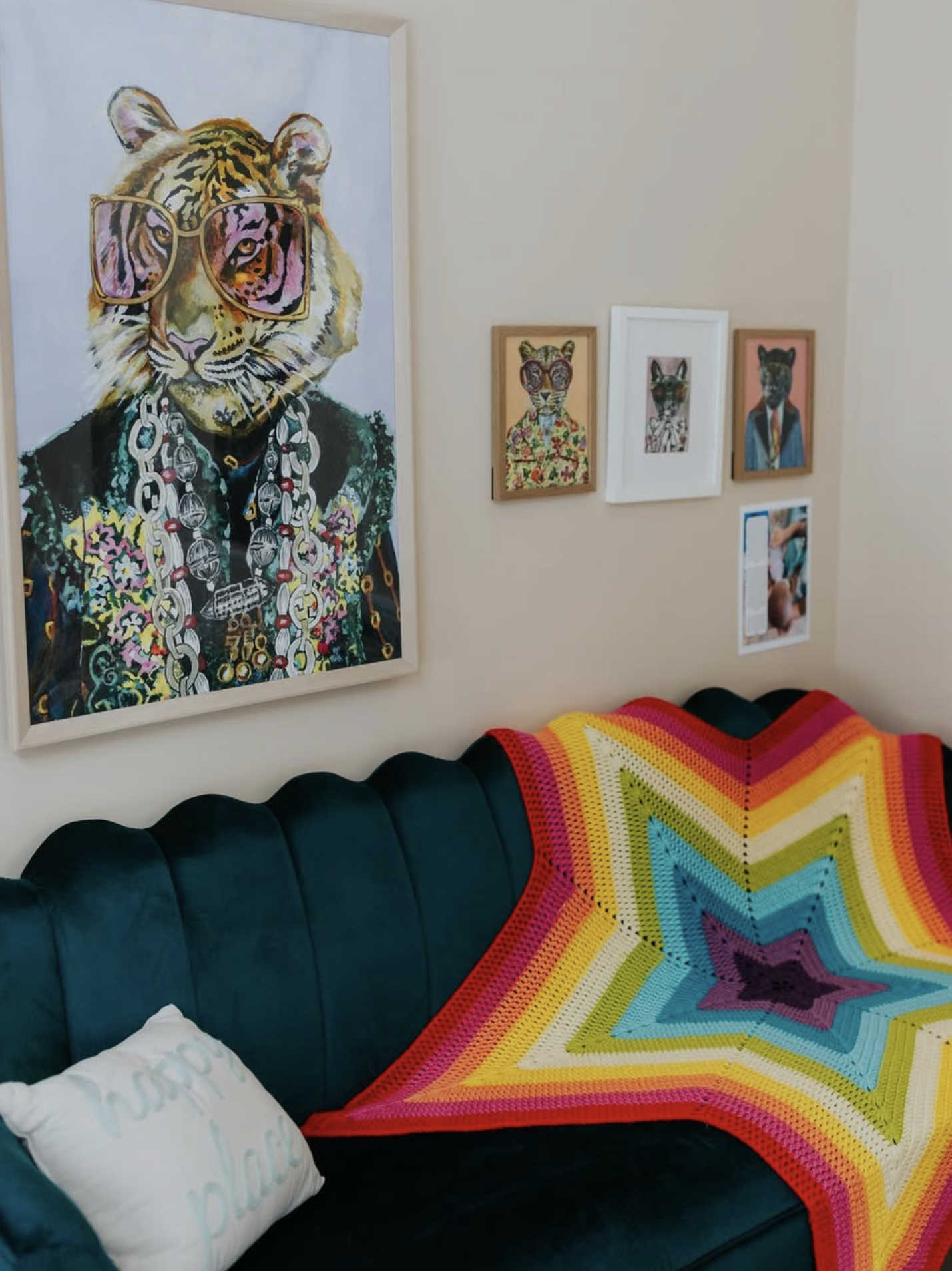 A living room corner with a dark green velvet couch covered by a colorful rainbow star-shaped crocheted blanket. There is a white pillow with writing on it. Above the couch, there are five framed pictures of anthropomorphic animals, including a tiger in sunglasses, dressed in colorful outfits, with the tiger also wearing large jewelry and chains.