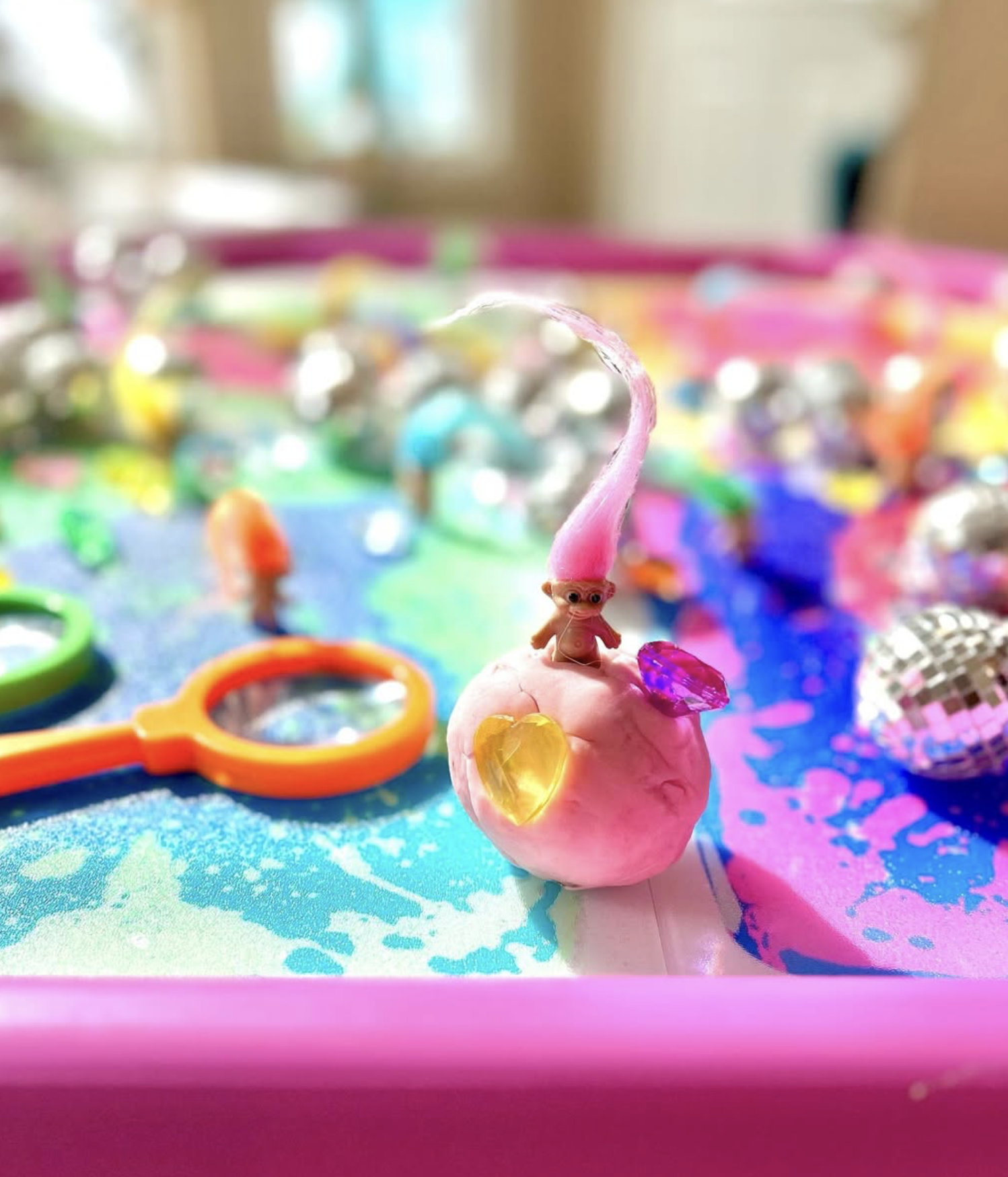 Colorful sensory bin with a small pink squishy toy, decorated with shiny heart-shaped stickers and a tiny plastic figure, surrounded by small mirrors, colorful rings, and glittery objects.
