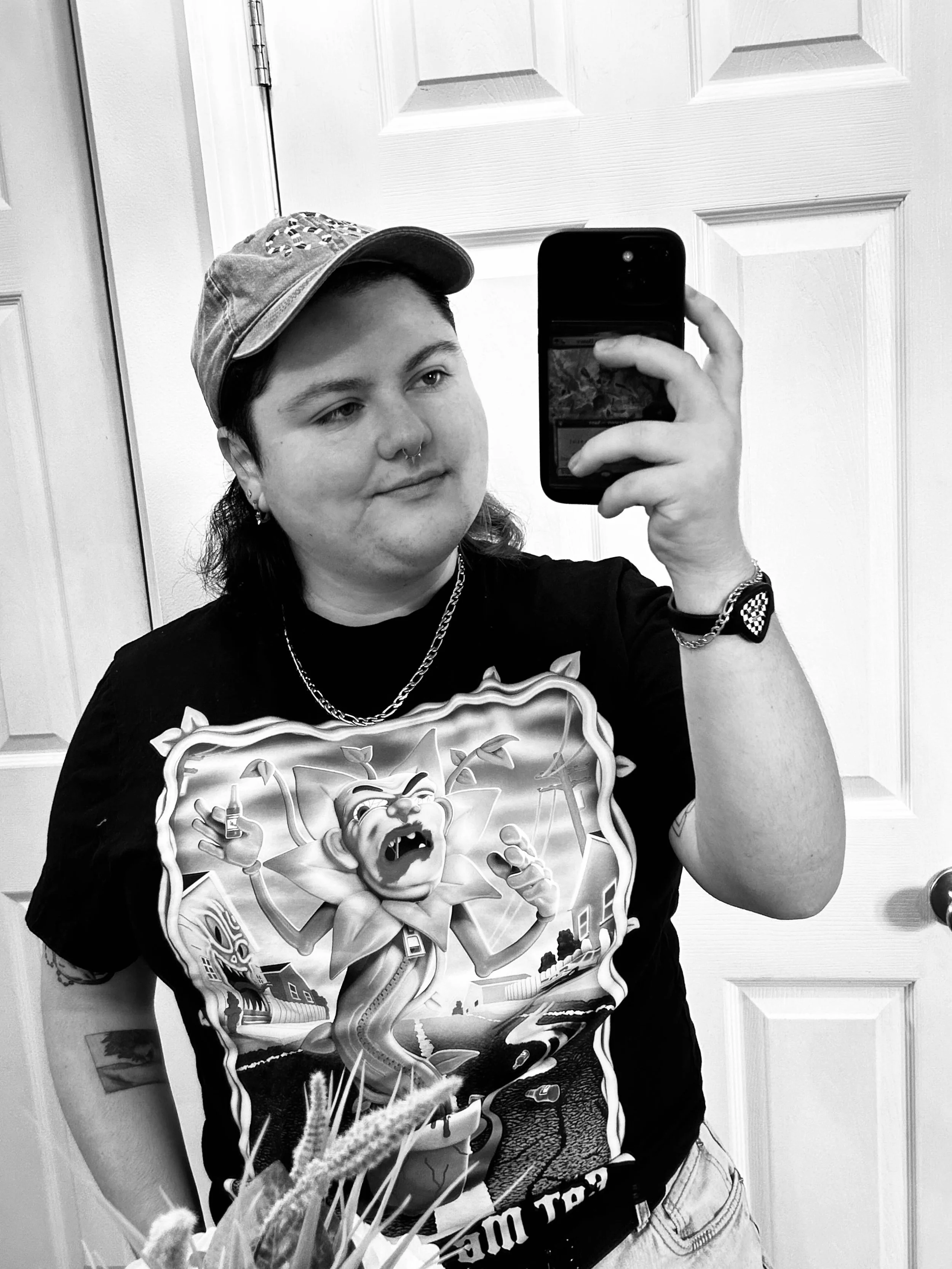 A person taking a mirror selfie in black and white, wearing a baseball cap, a graphic T-shirt with a cartoon character, and accessories like earrings, a necklace, and a bracelet.