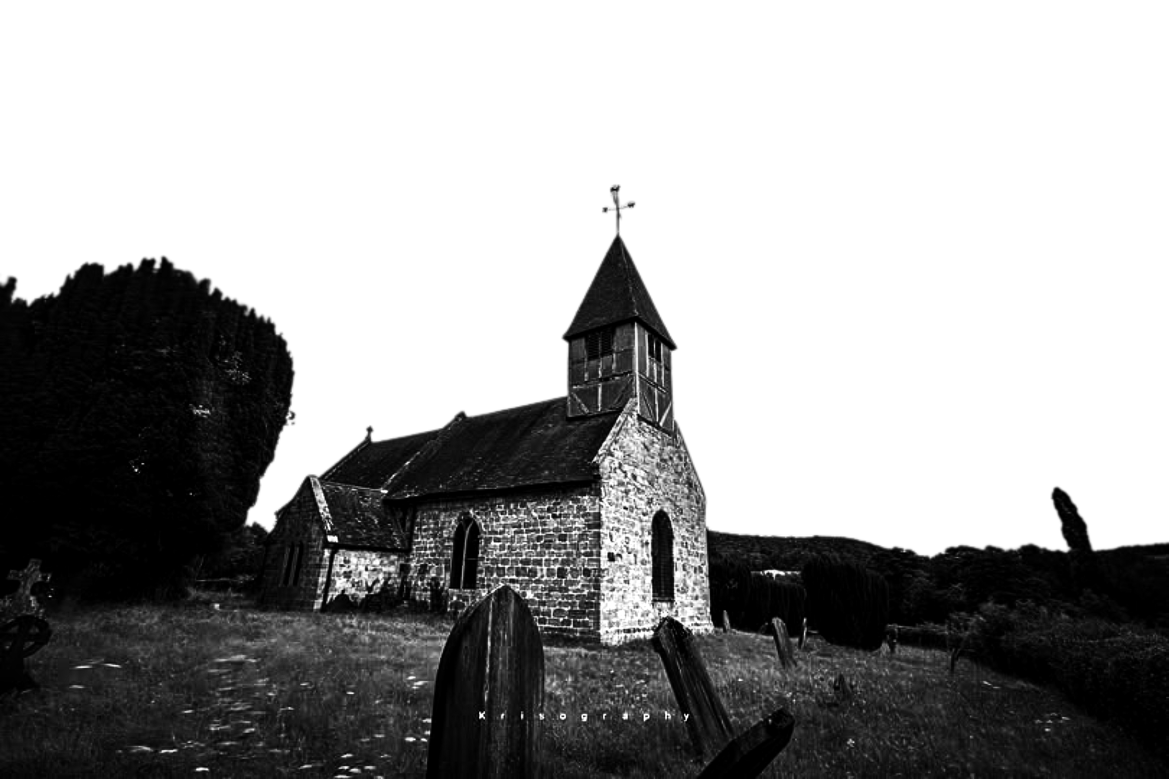 A black and white photo of an old stone church with a tall steeple, surrounded by a graveyard with weathered tombstones and plants.