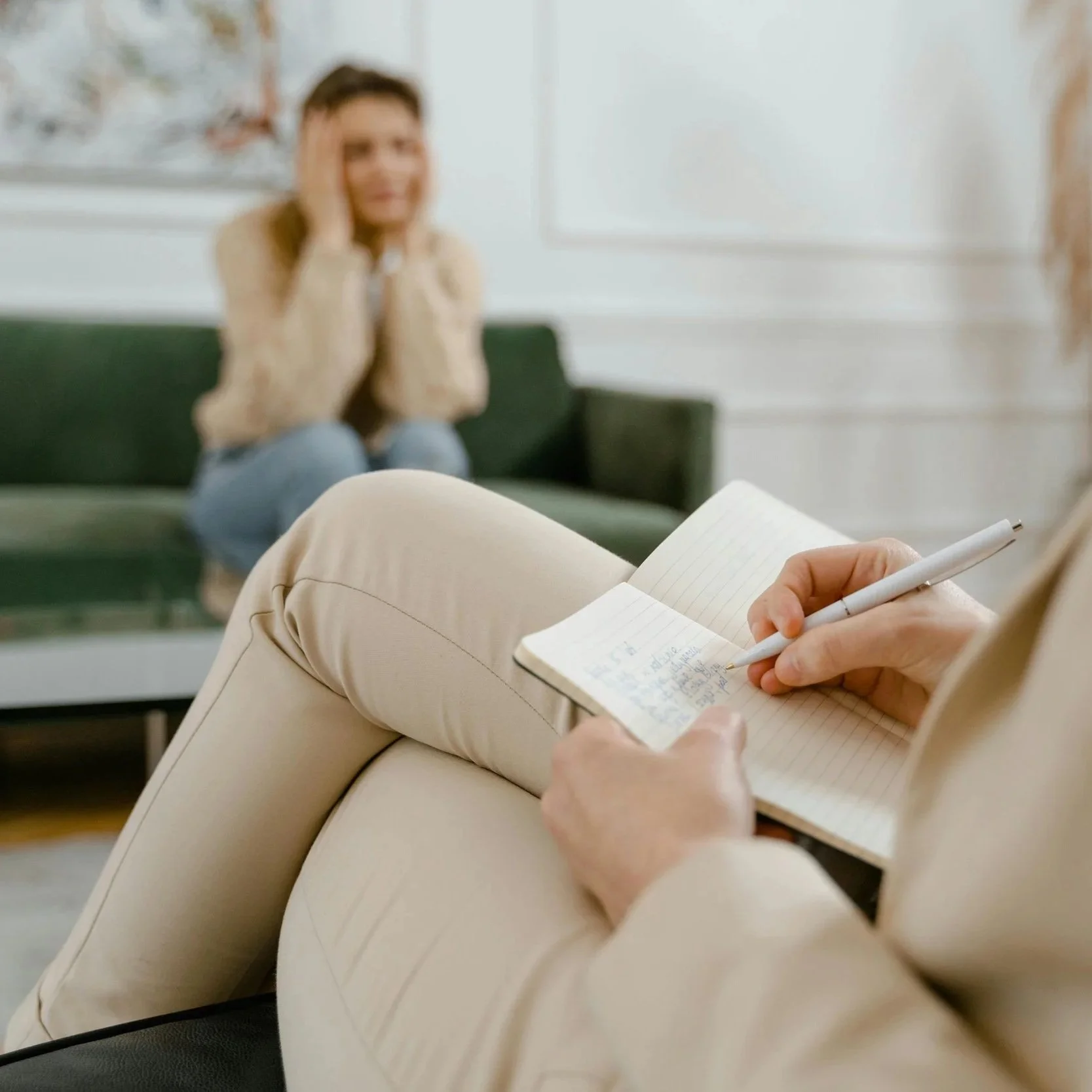 A therapist taking notes while a woman in a beige sweater sits on a green couch in the background, appearing distressed.