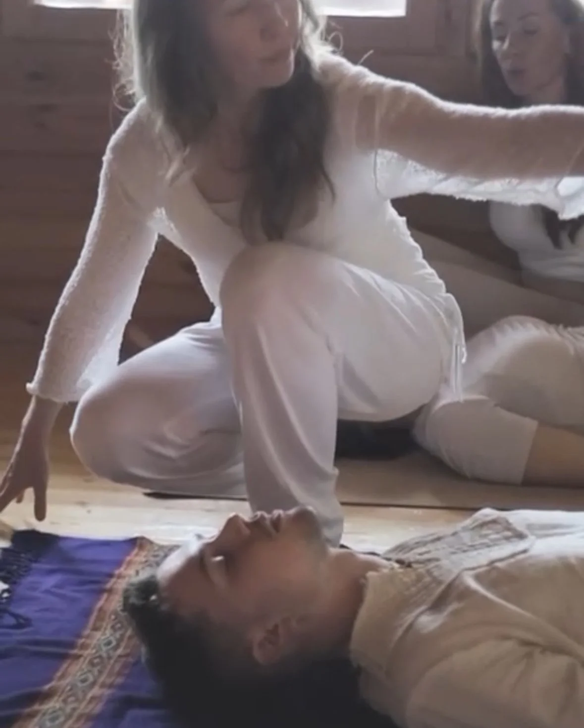 A woman with long brown hair in white clothing is kneeling on the floor, leaning over a man lying on a rug, with her hand held above his face. Another woman is sitting behind her, watching.