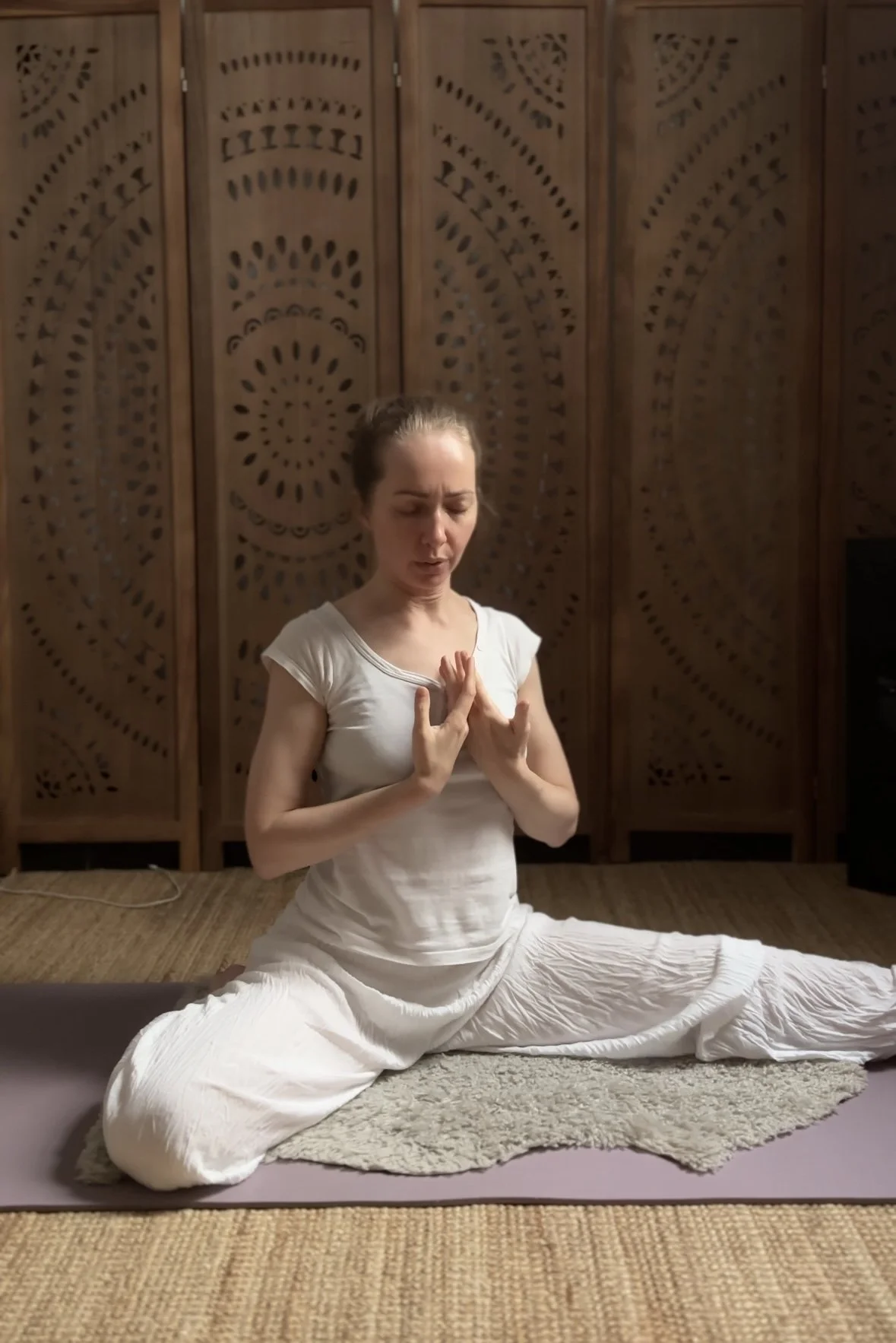 A woman practicing yoga on a purple mat with a small gray rug underneath, in a room with wooden lattice walls, dressed in white, sitting in a split position with her hands in a prayer position against her chest and eyes closed.