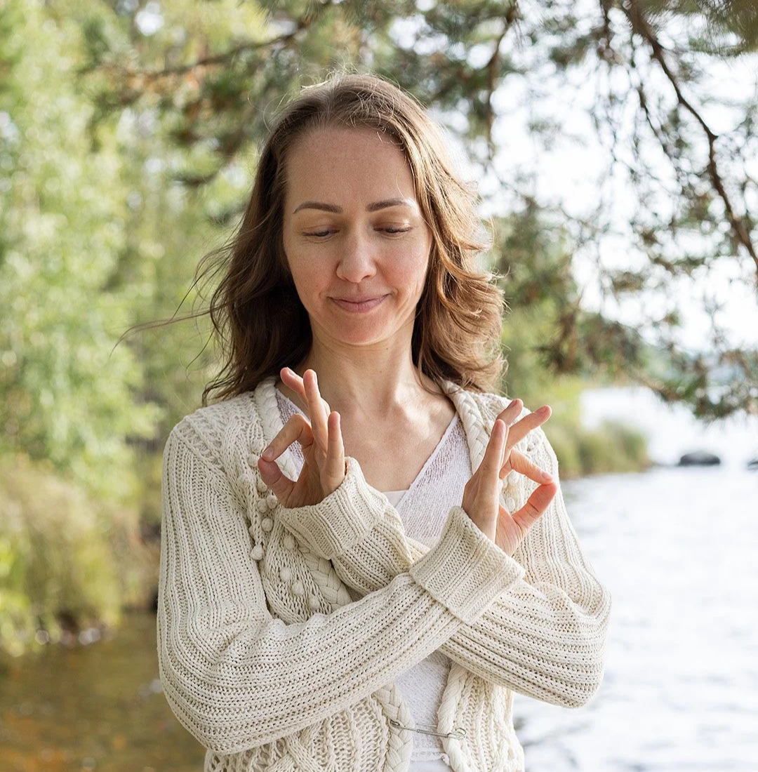 Woman standing outdoors near water, making a calming gesture with her hands, wearing a cream-colored knitted sweater, with trees and water in the background.