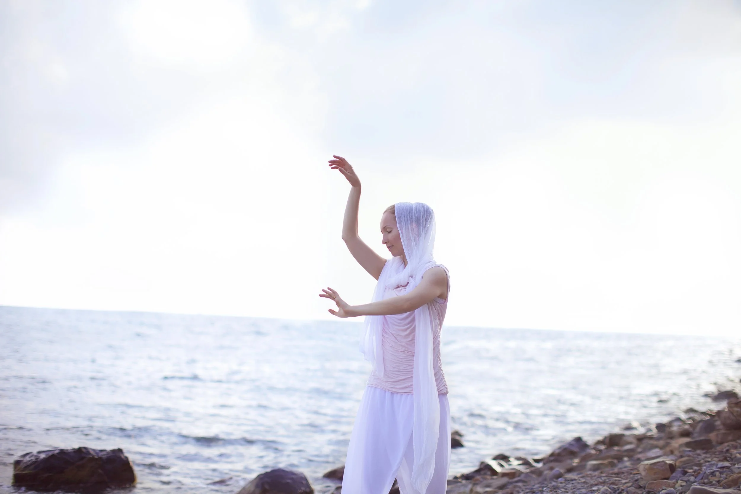 Woman in flowing white clothing and a sheer headscarf standing on a rocky beach near the water, with arms raised and eyes closed.