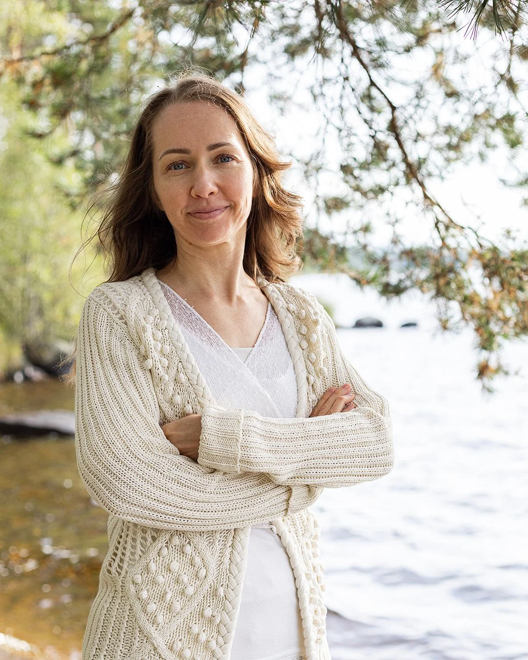 Woman in cream-colored knit cardigan standing outdoors by a body of water, with trees and rocks in the background.