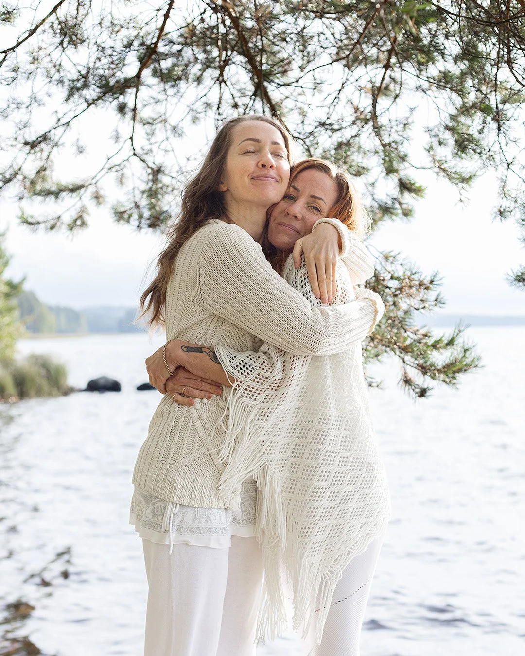 Two women with light skin and similar hair color hugging each other outdoors near a body of water under a tree.