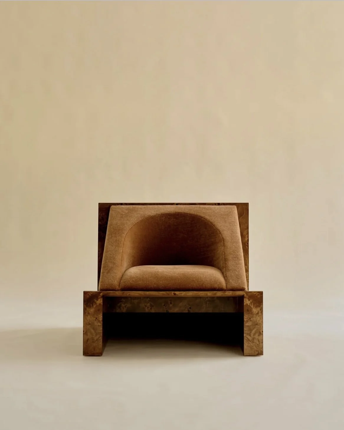 A modern single armchair with a rounded back in a soft brown fabric, placed on a wooden platform against a plain beige wall.