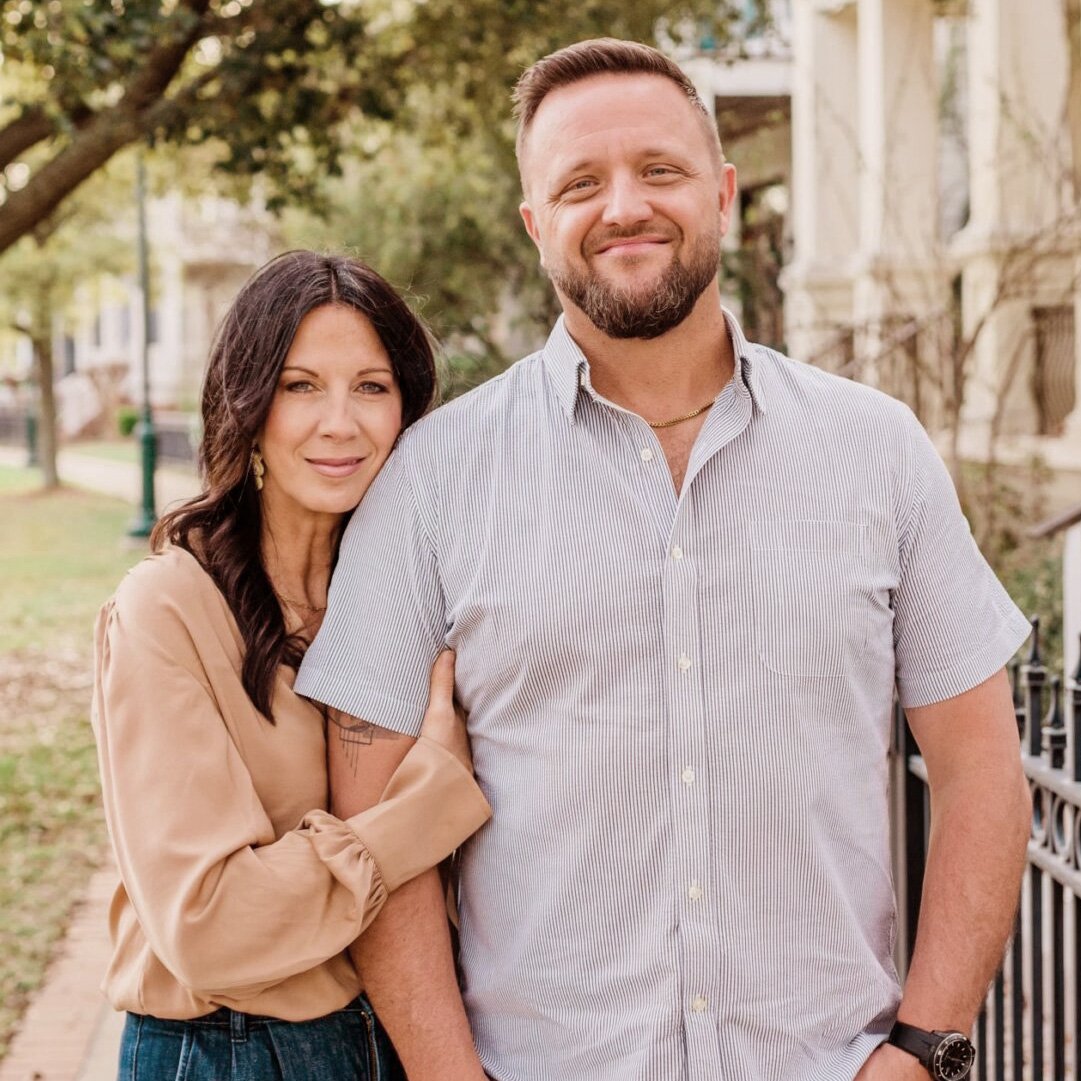 Couple standing outdoors, smiling, with the woman holding the man’s arm