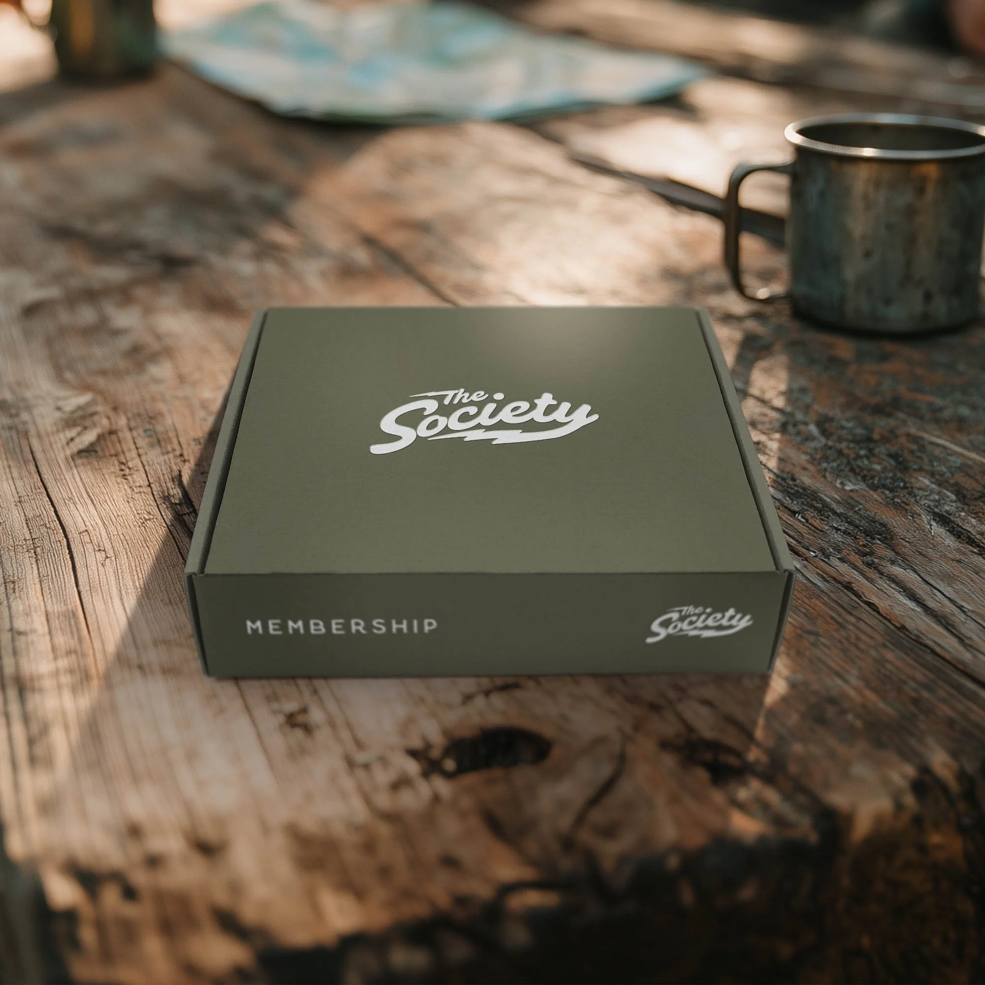 Green “The Society” box on a rustic wooden table beside a metal cup.