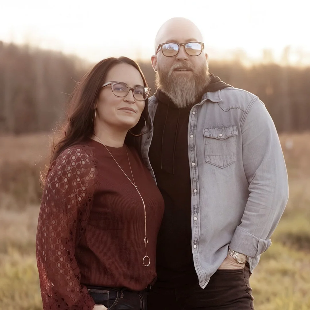 Two adults standing outdoors at sunset, wearing glasses and casual clothes, in a wooded setting.