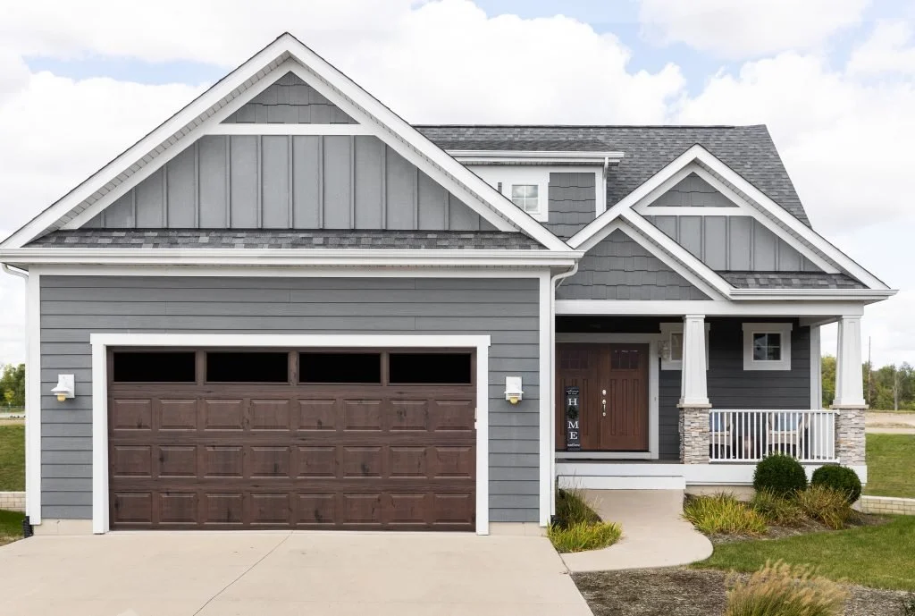Front view of a modern gray house with a brown garage door, front porch with white railing, and landscaped yard.