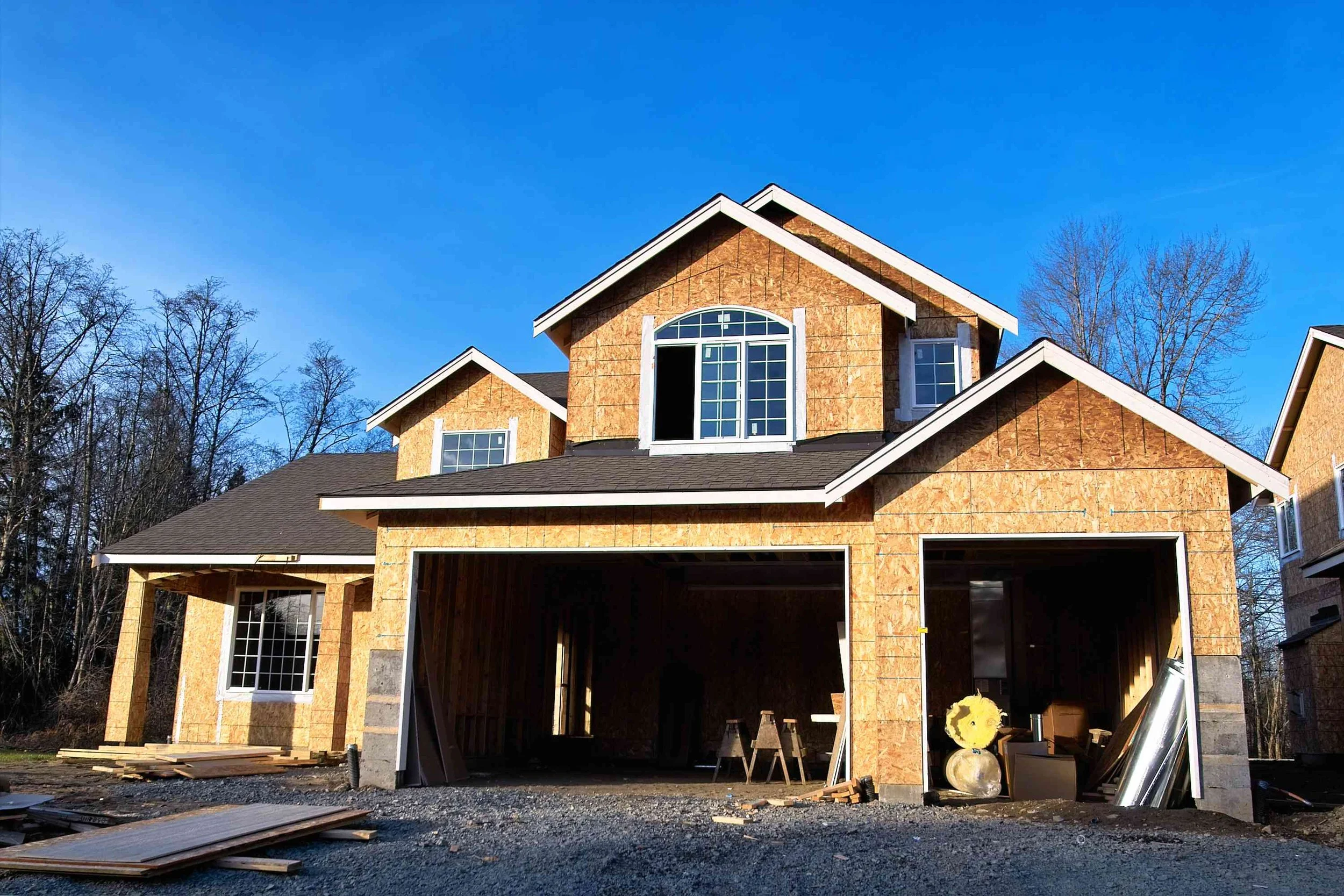 Under construction two-story house with unfinished exterior, open garage, construction materials inside and outside, and a blue sky. new construction garage door. contractor