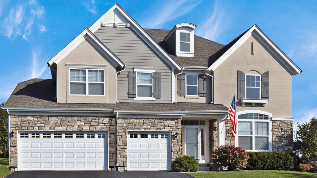A large, two-story suburban house with stone and beige siding exterior, featuring front-facing garage doors, multiple windows with shutters, and an American flag hanging near the entrance. Patriotic Garage door company