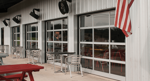 Empty outdoor seating area with metal tables and chairs in front of a building with large glass windows, metal siding, and an American flag hanging. commercial garage doors