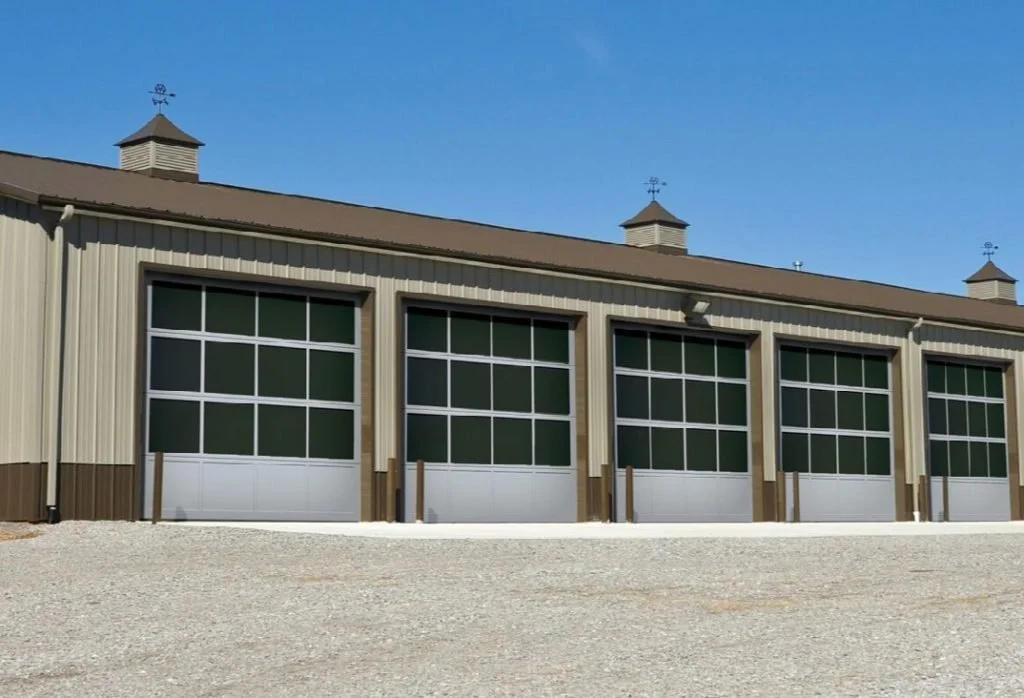 Large building with multiple garage doors, metal siding, and a brown roof with three small cupolas topped with weather vanes, under a clear blue sky.