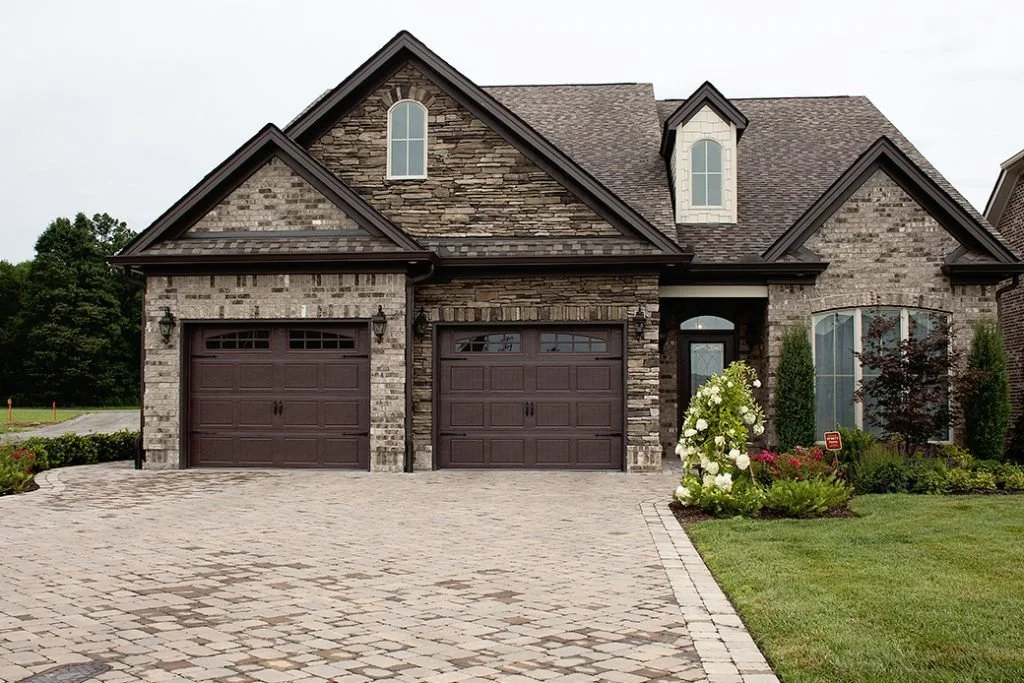 A house with brick and stone exterior, two brown garage doors, a driveway, and landscaped front yard with green grass, bushes, and flowers. upgraded garage doors