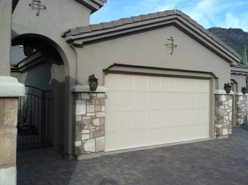 A residential garage with a closed cream-colored door, stone and stucco walls, and outdoor wall-mounted lanterns. There is a gated side passage to the left. wind load garage door