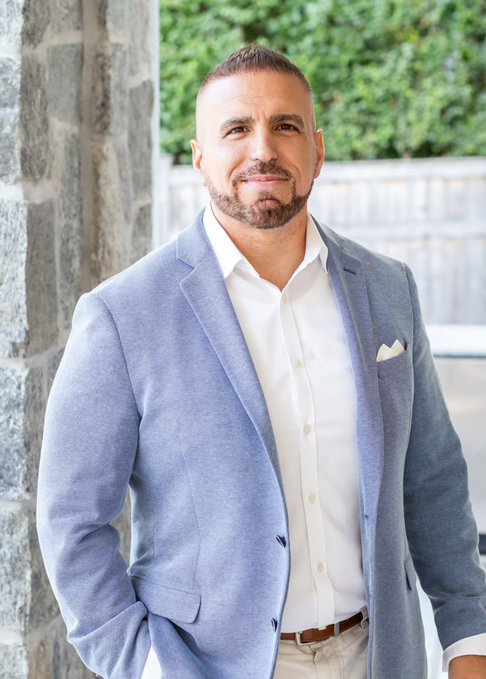 Portrait of a man in a light blue blazer and white shirt standing outdoors near stone wall and greenery.