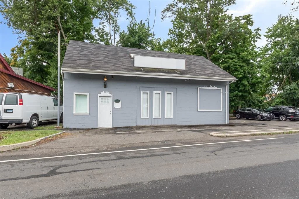 A small, gray building with a sloped roof and boarded-up windows, located on a street with parked cars and trees in the background.