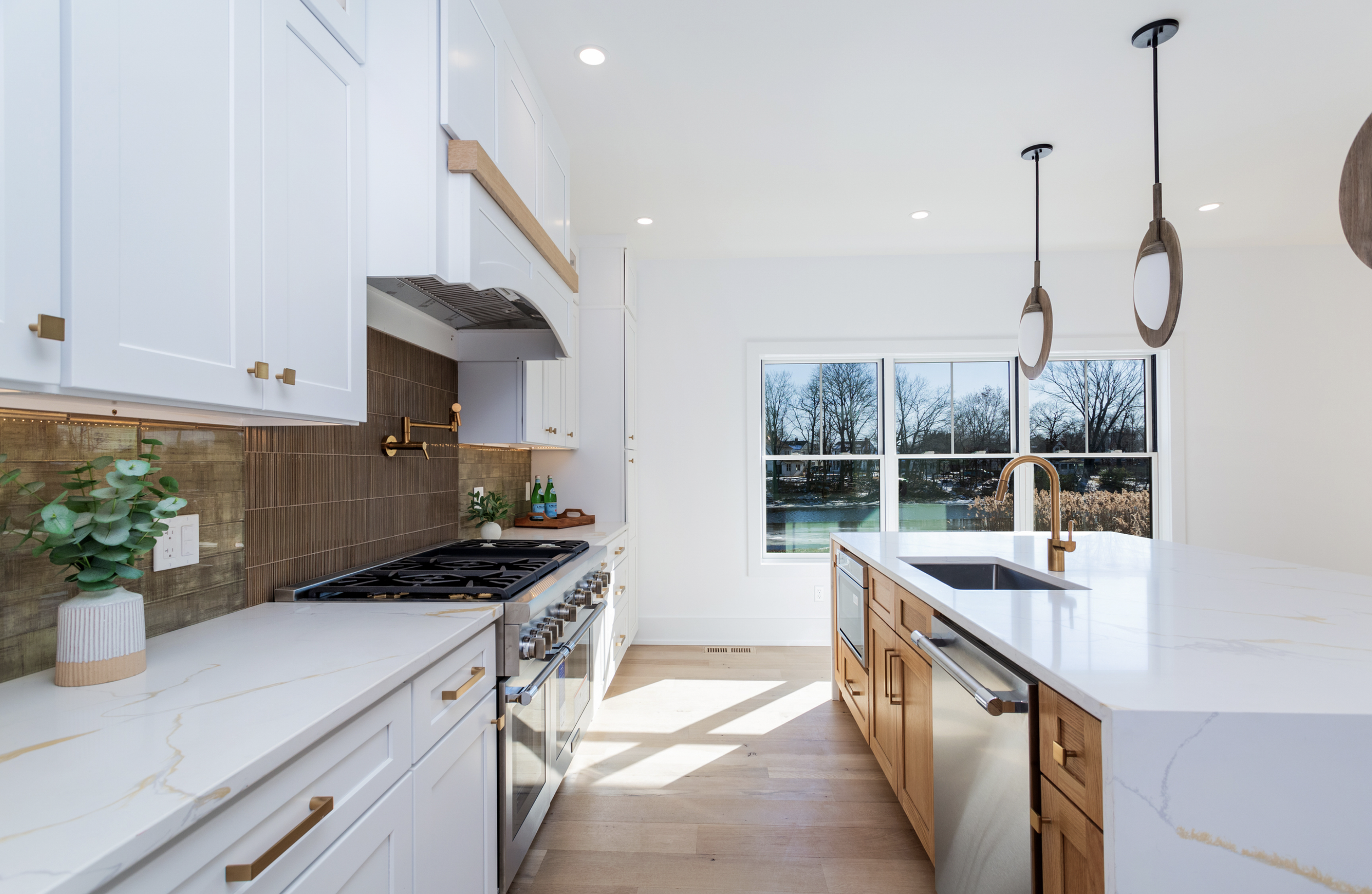Modern kitchen with white cabinets, wooden accents, a large white marble countertop island with a black sink, gold fixtures, a glass window overlooking a lake with trees, and pendant lights.