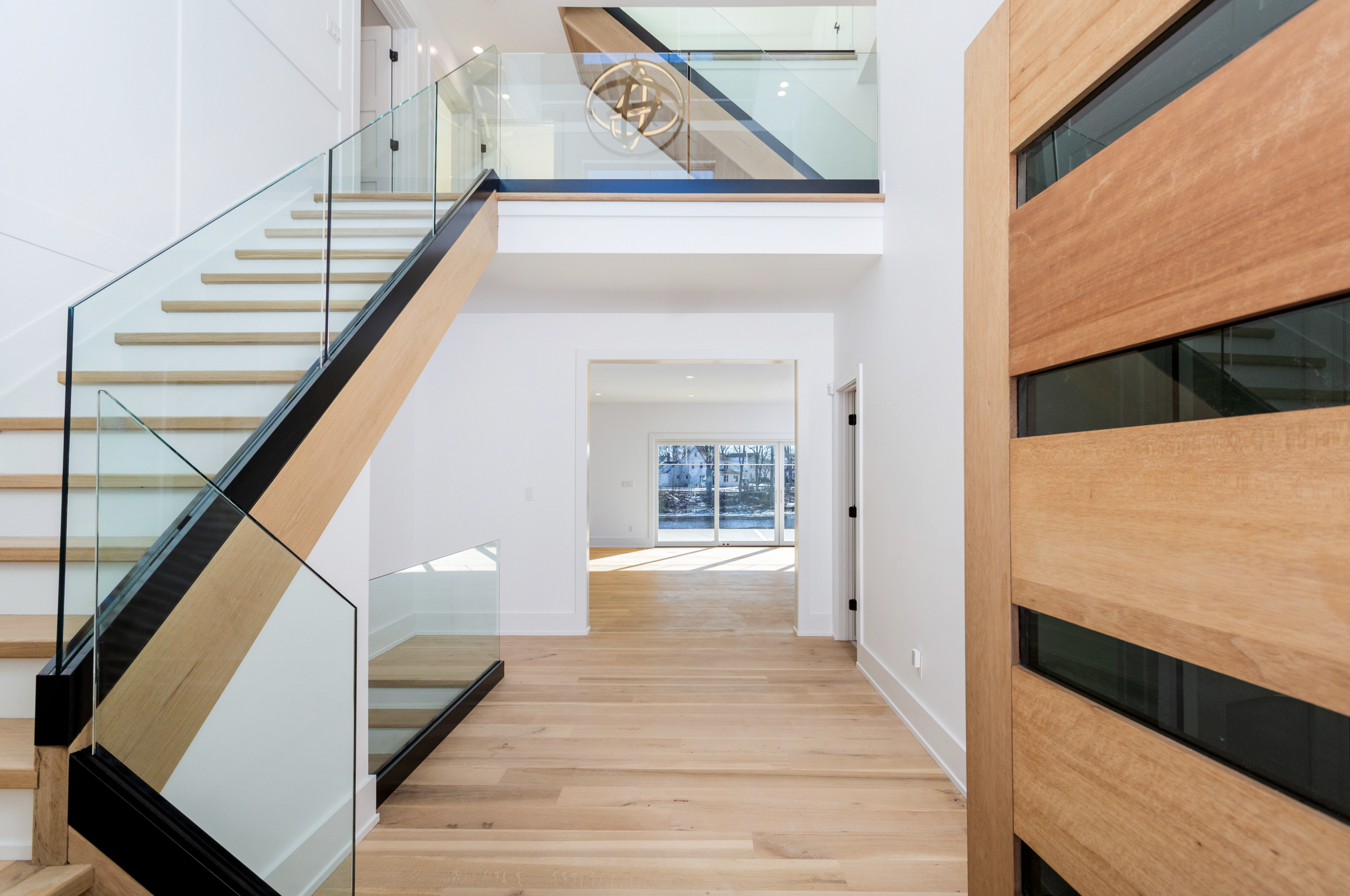 Interior view of a modern house with a staircase, glass railing, light wooden floors, and a wooden door with horizontal glass panels leading to a bright room with large windows.