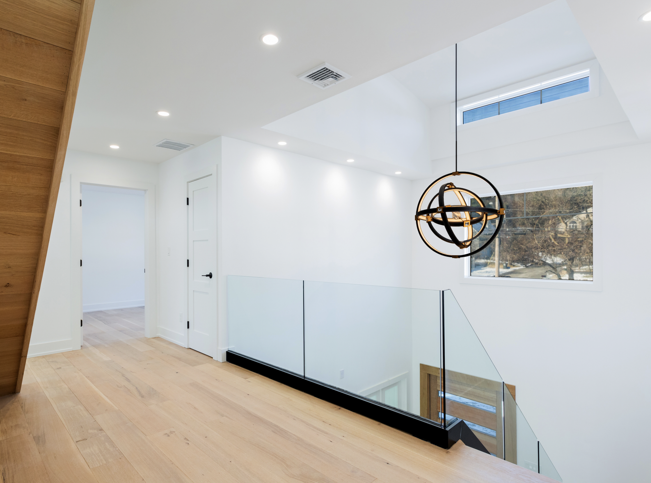 Modern interior space with white walls, light wood flooring, a black and gold spherical pendant light, large windows, and a glass railing beside a staircase.