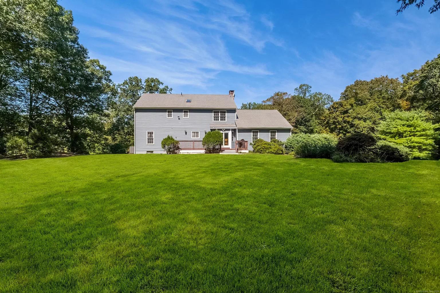 A large gray house with multiple windows and a small deck is situated on a lush green lawn with trees in the background, under a blue sky with few clouds.