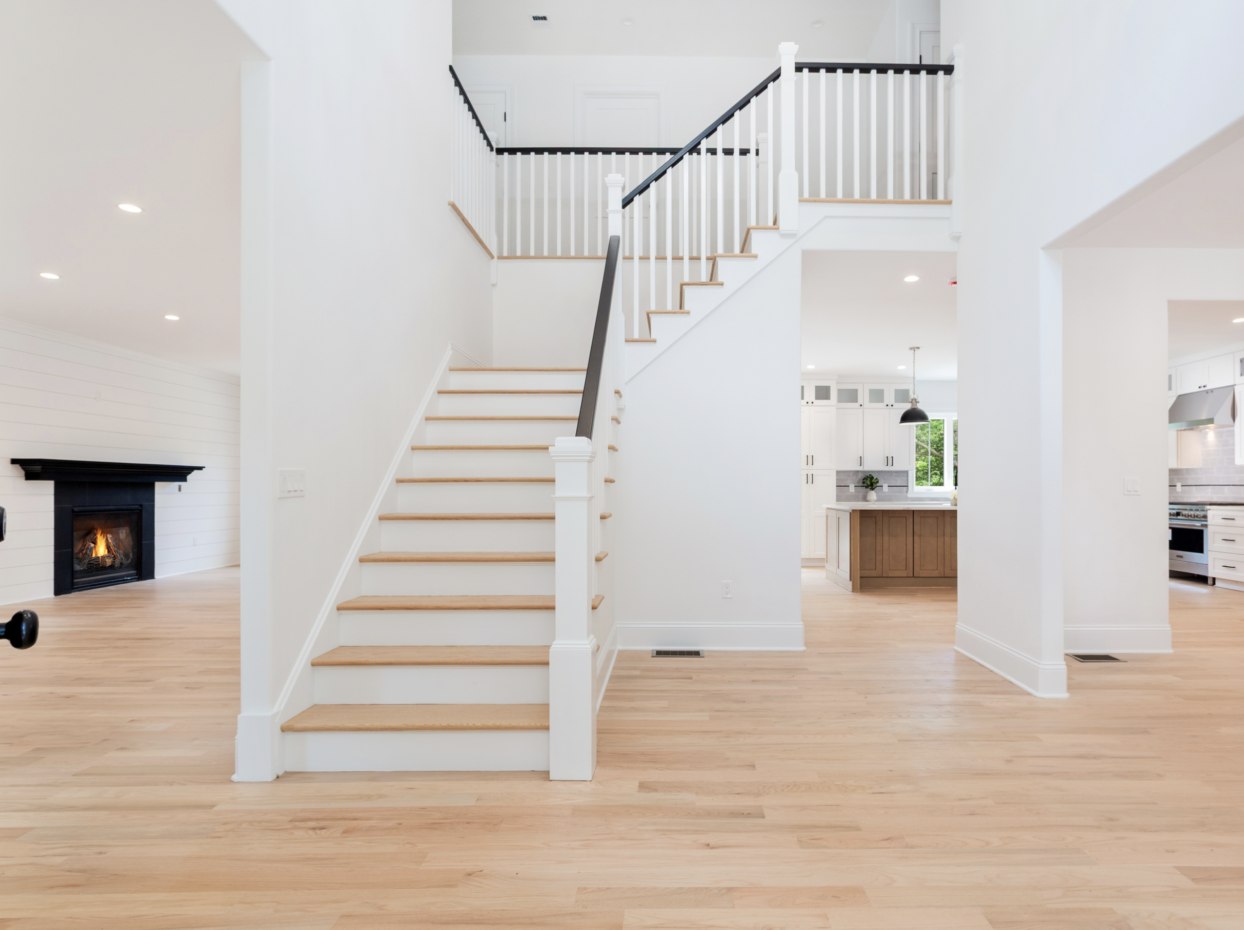 Interior of a modern home with wooden floors, a central staircase with white balusters and a black handrail, open kitchen with white cabinets and a wooden island, and a fireplace in the living area.