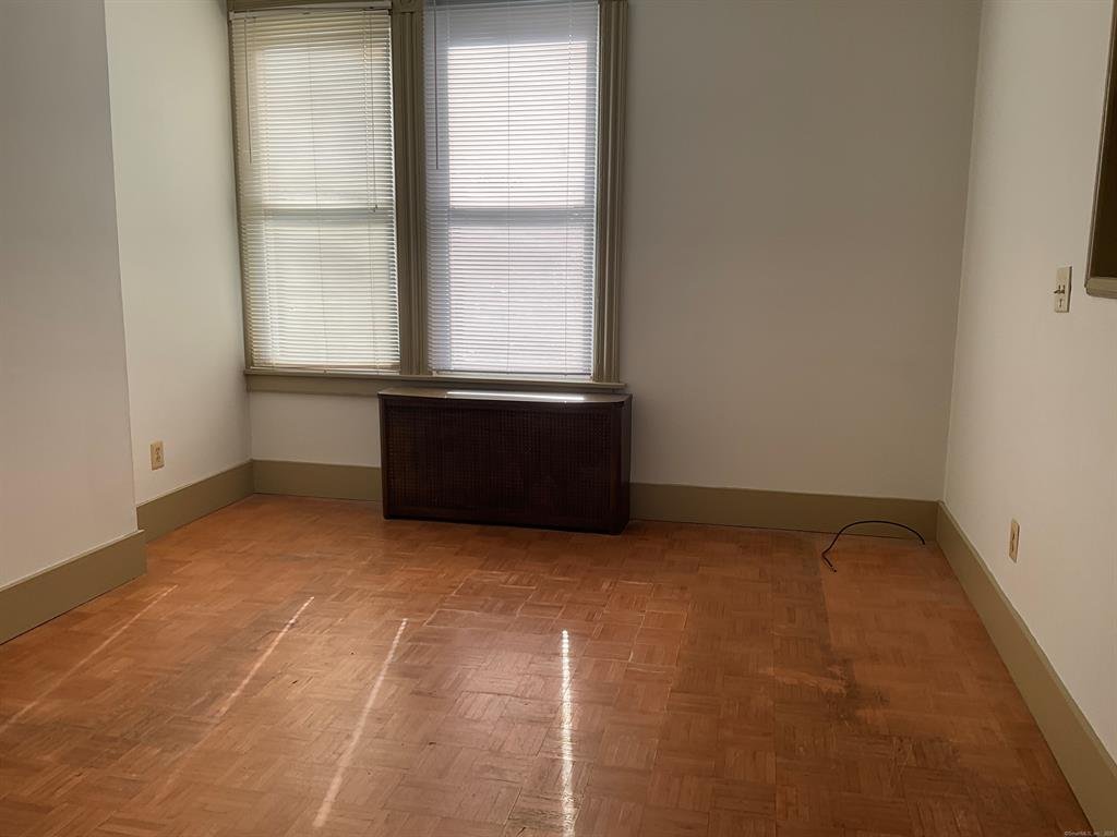 Empty room with hardwood floors, beige baseboards, white walls, and a window with blinds. There is a heater or radiator in front of the window and a visible electrical cord on the floor.