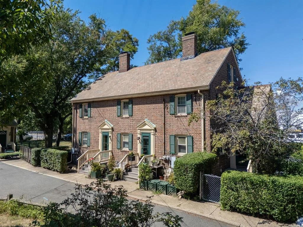 A two-story brick house with green shutters, front porch steps, and potted plants, surrounded by trees and shrubbery.
