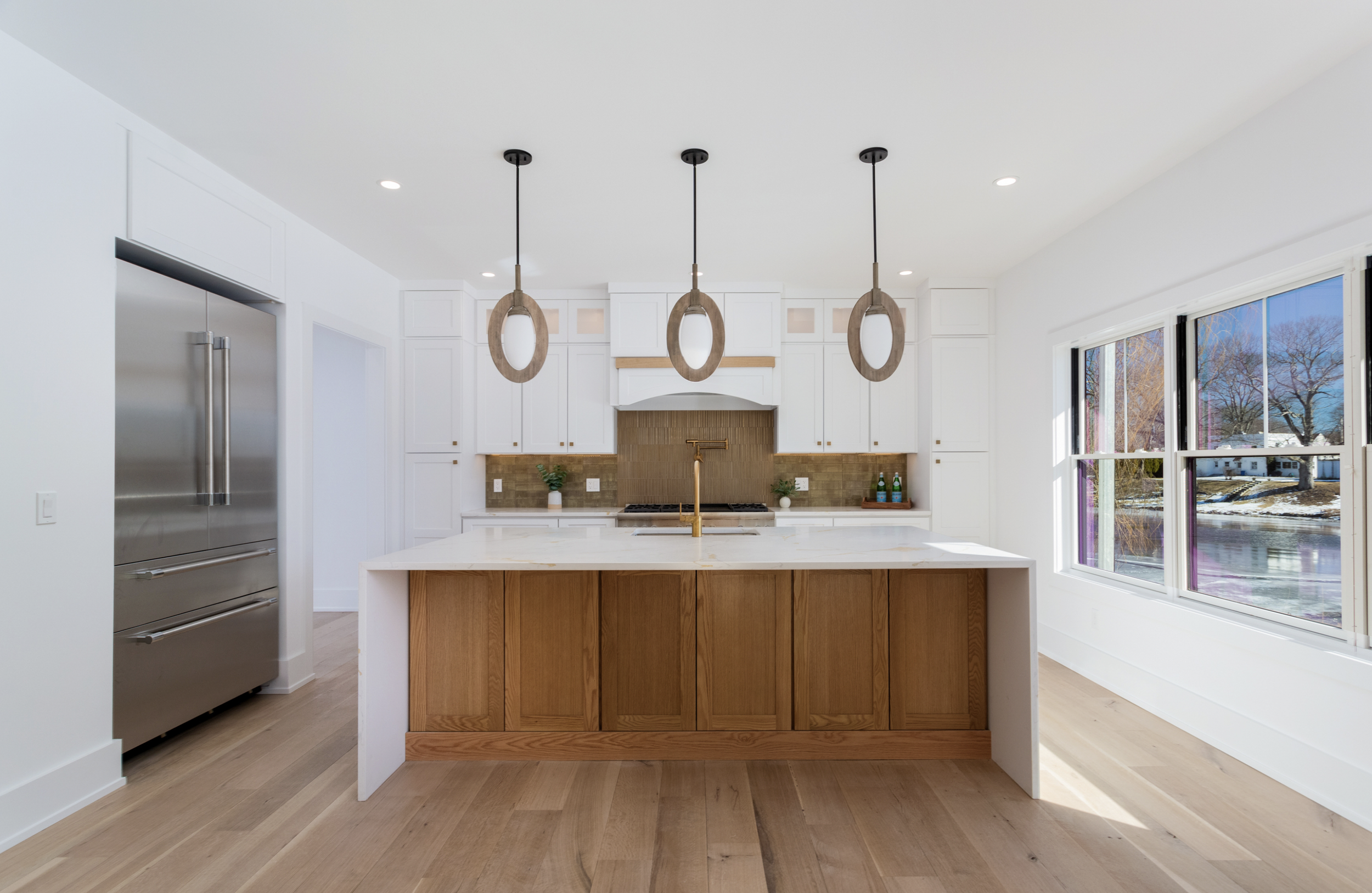 Modern kitchen with white cabinets, wooden island, pendant lighting, large windows showing outdoor view, hardwood floors, stainless steel refrigerator, and minimal decor.