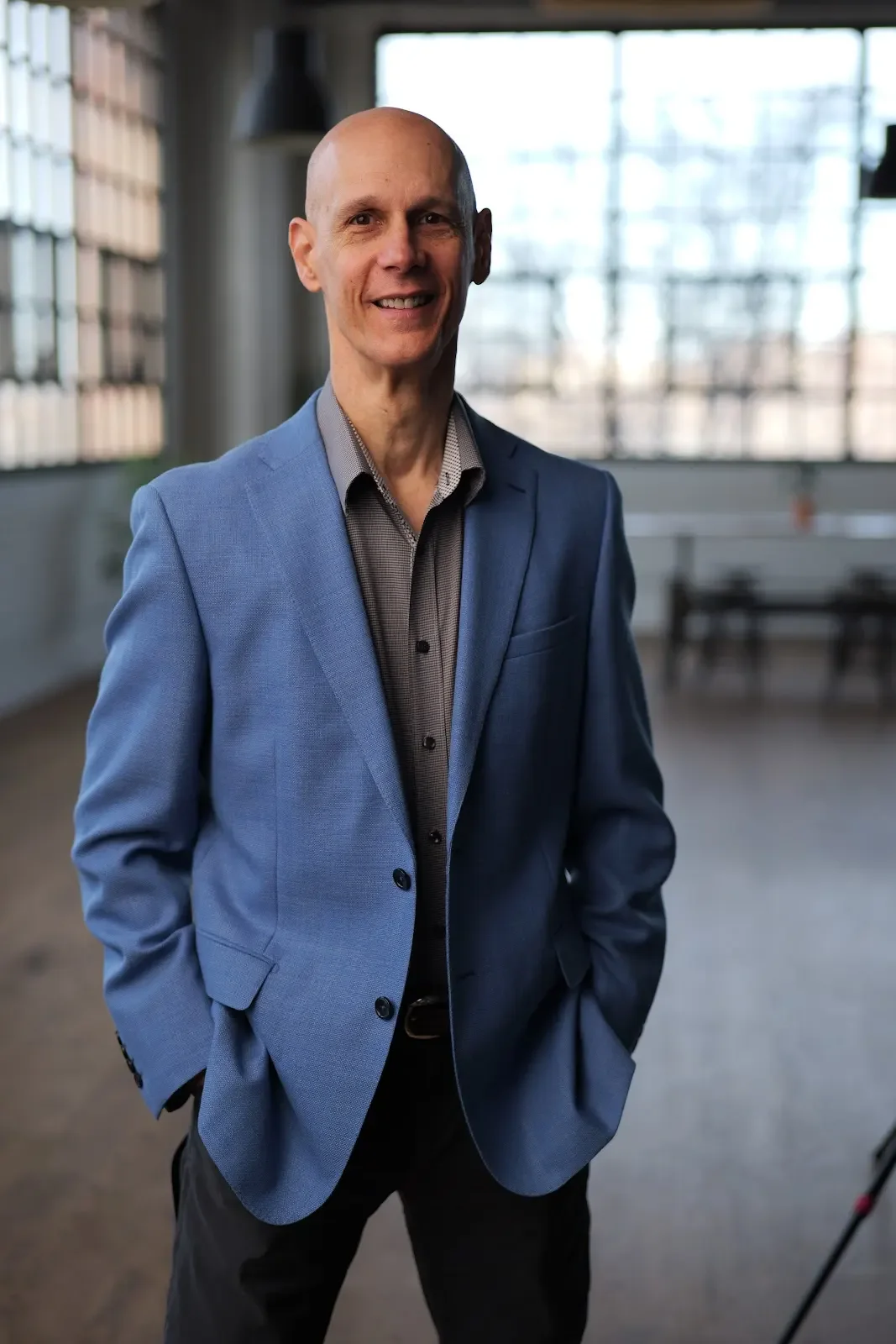 Man in blue suit jacket smiling, standing indoors with large windows in the background.
