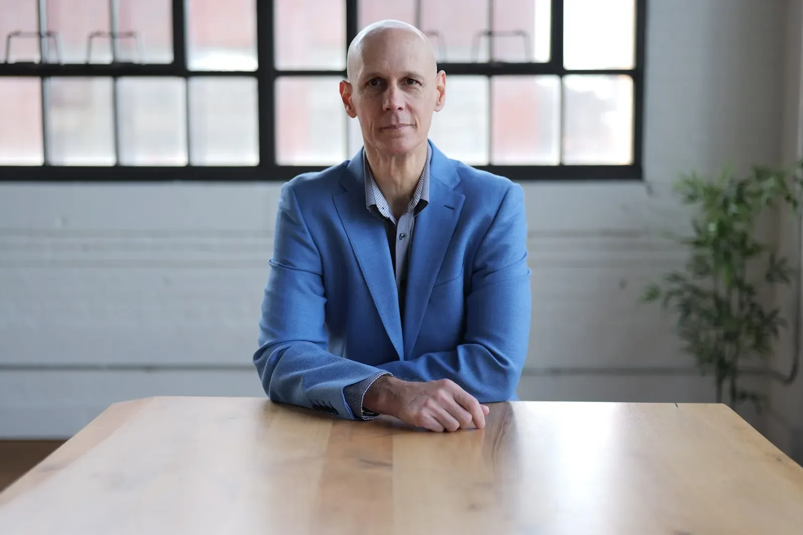 A bald man in a blue blazer sitting at a wooden table in a room with large windows and a potted plant in the background.