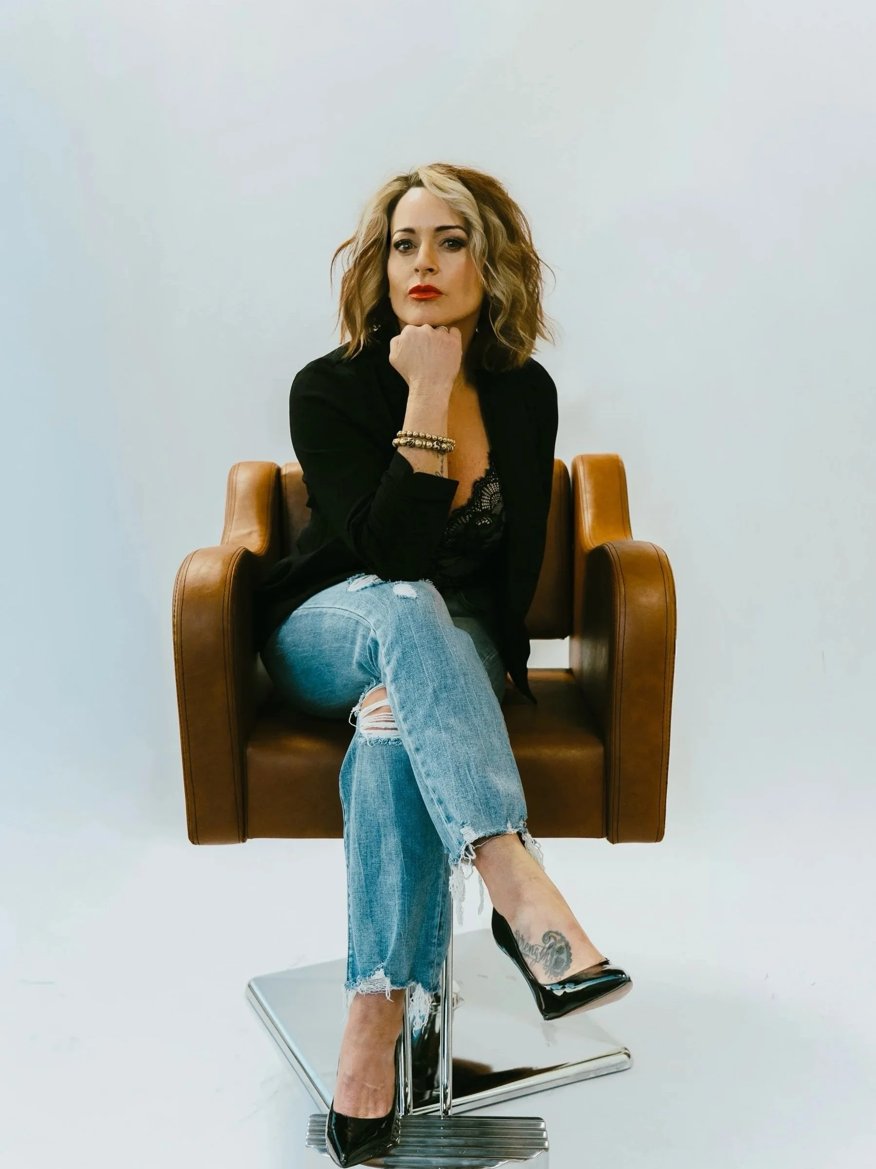 A woman with blonde, wavy hair sitting on a brown leather chair against a plain white background. She is dressed in a black jacket, lace top, ripped jeans, and black high heels, with one hand resting under her chin and a confident expression.