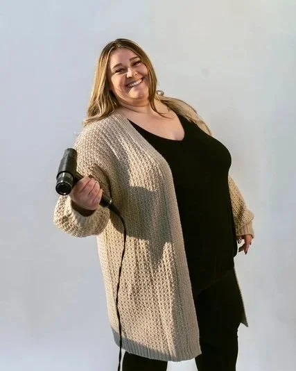 A woman with long blonde hair smiling and holding a curling iron in her right hand, wearing a beige cardigan over a black top, standing against a plain white background.